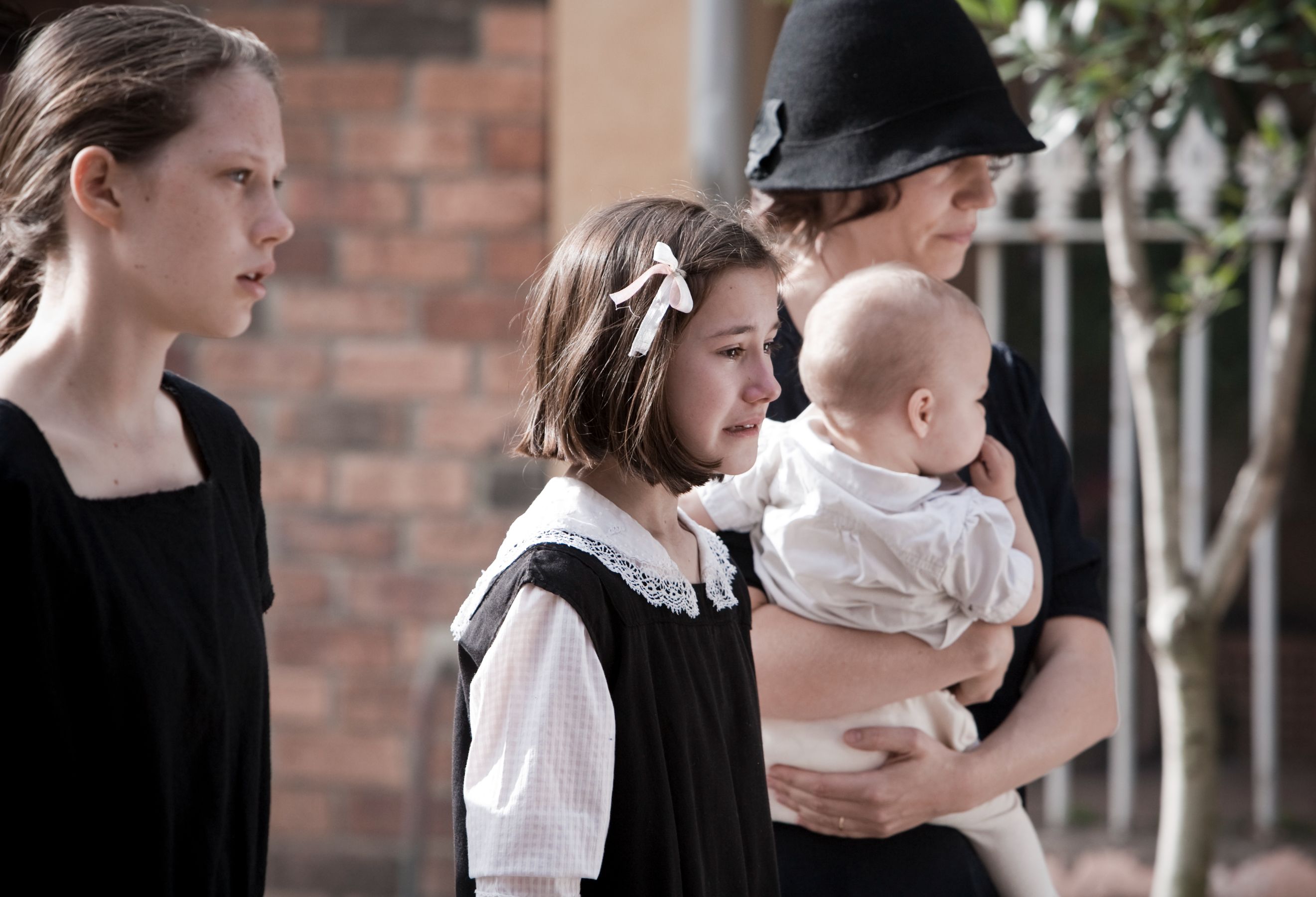 Two girls in black dresses walk solemnly, one with a white bow in her hair, showing sadness on their faces. A woman in the background holds a baby, also dressed in white, while behind them stands a brick wall and greenery.