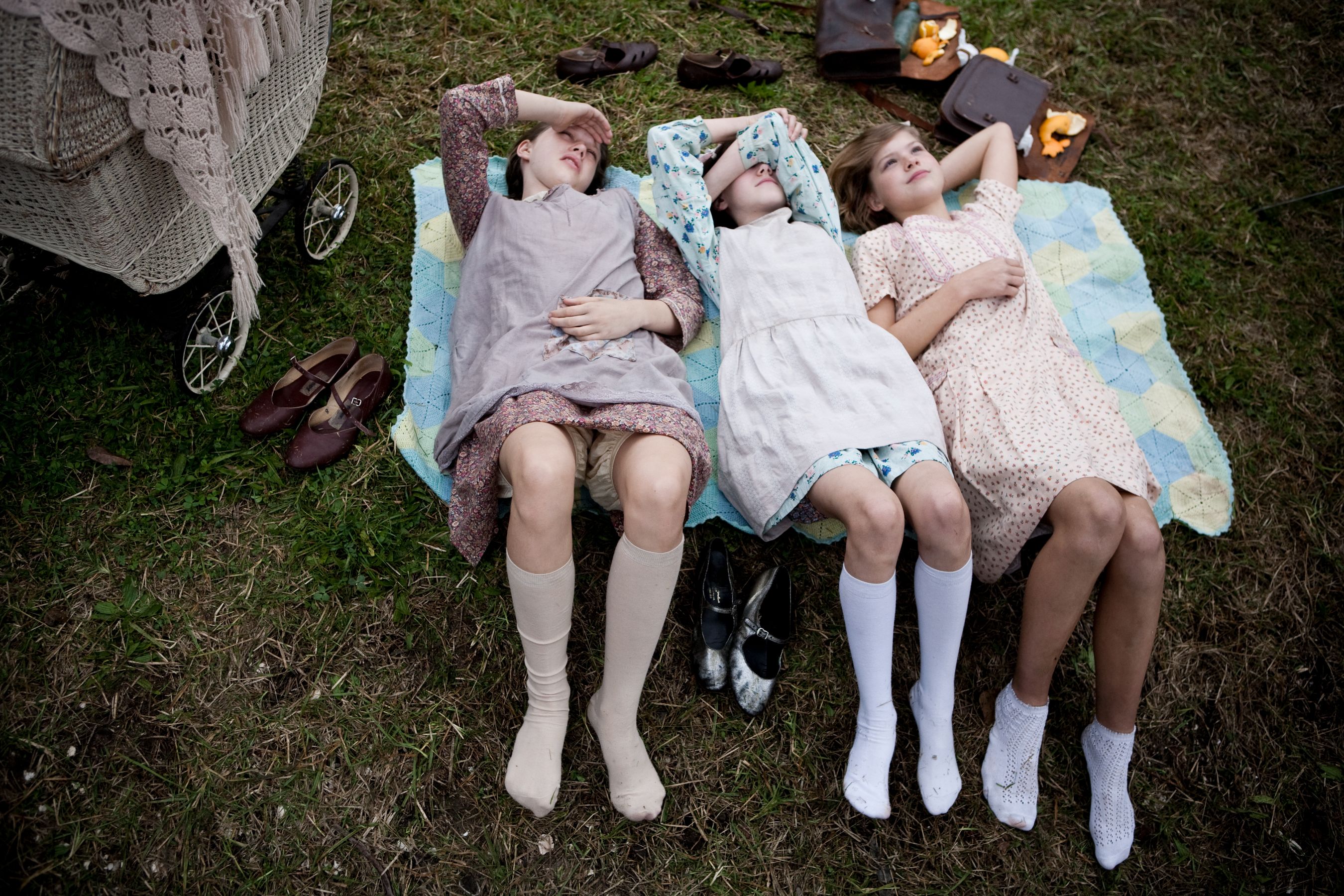 Three girls lay on a quilt in a grassy area, shielding their eyes from the sun. They each wear different dresses and knee-high socks, with shoes scattered around them, along with a baby stroller and bags in the background.