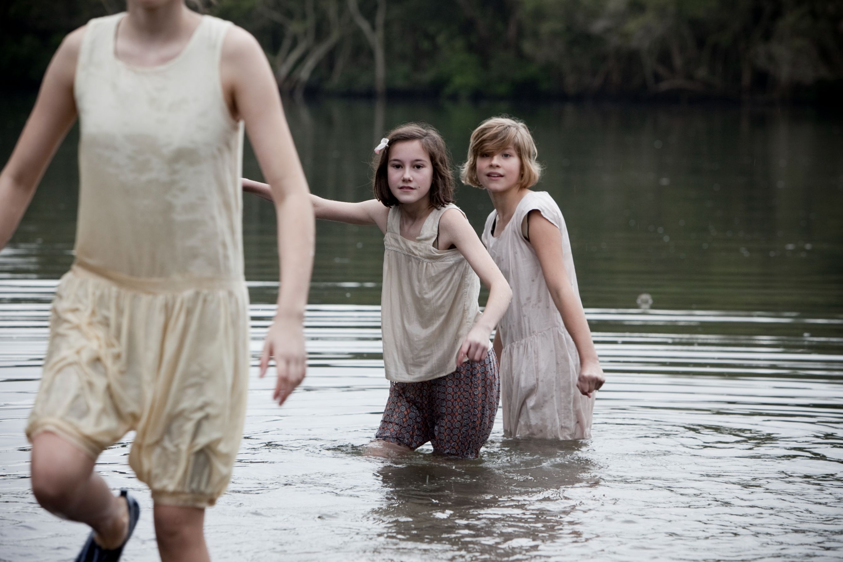 Two girls wade through shallow water, with one reaching out toward the other. They wear light-colored, loose-fitting clothing, and the surroundings feature lush vegetation reflected in the water's surface. The atmosphere appears calm and subdued.