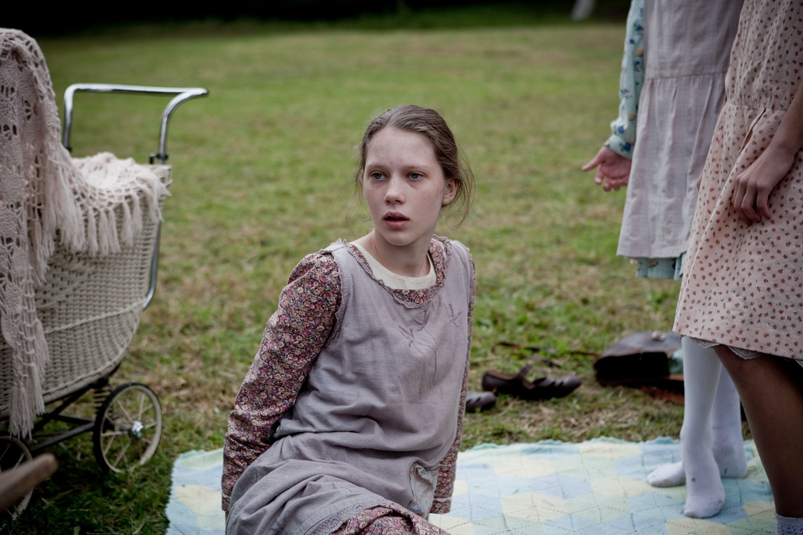 A young girl sits on a blanket in a grassy field, looking surprised or concerned. She wears a grey dress with a floral pattern underneath and appears to be in a moment of tension, while other children stand nearby. A baby carriage is partially visible in the background.