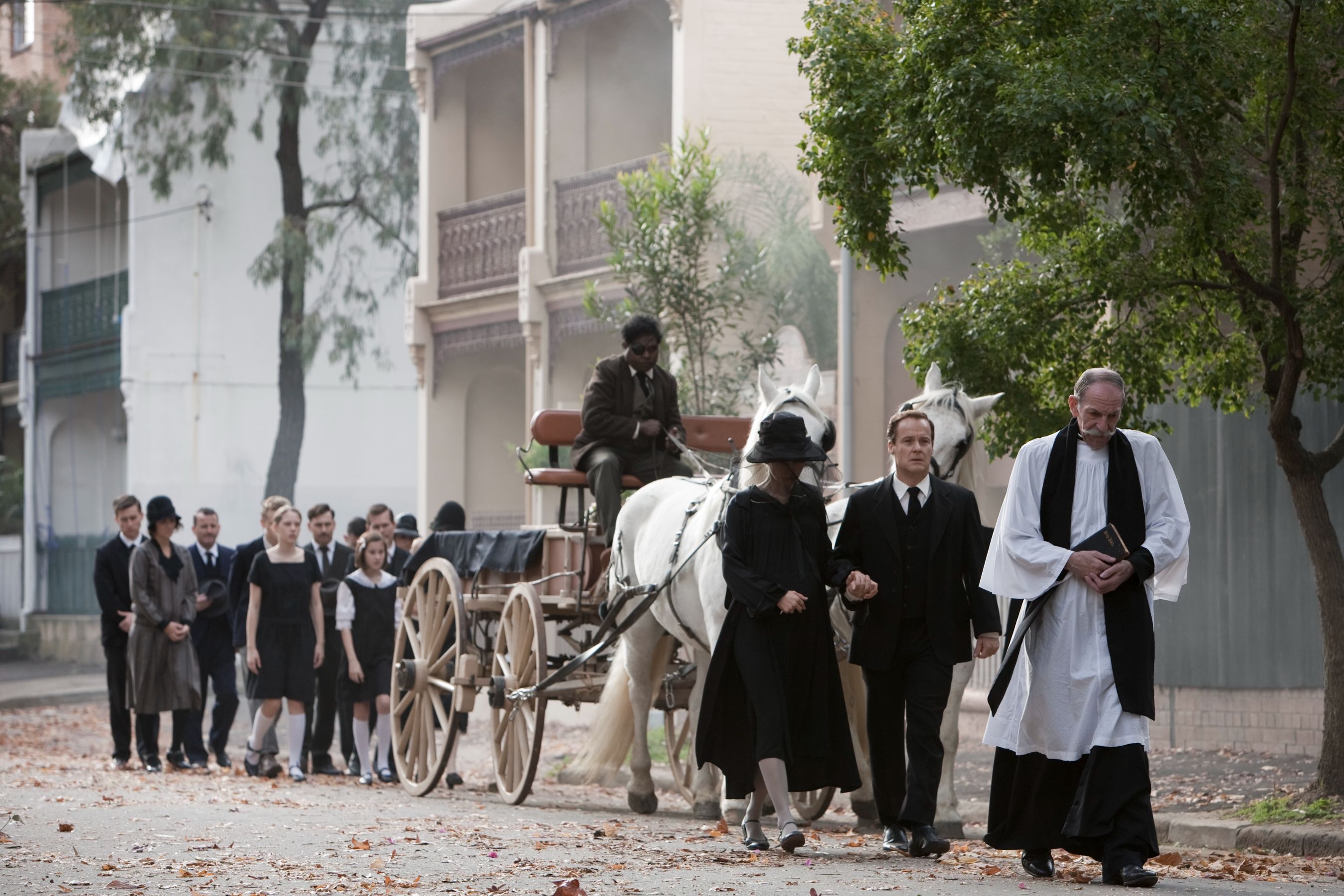 A group of mourners dressed in black attire walks solemnly down a tree-lined street, following a horse-drawn carriage. Among them, a clergyman in white and black robes holds a book, while a woman in a black dress and hat walks hand-in-hand with a man in a suit. The atmosphere is somber, with fallen leaves scattered on the ground.