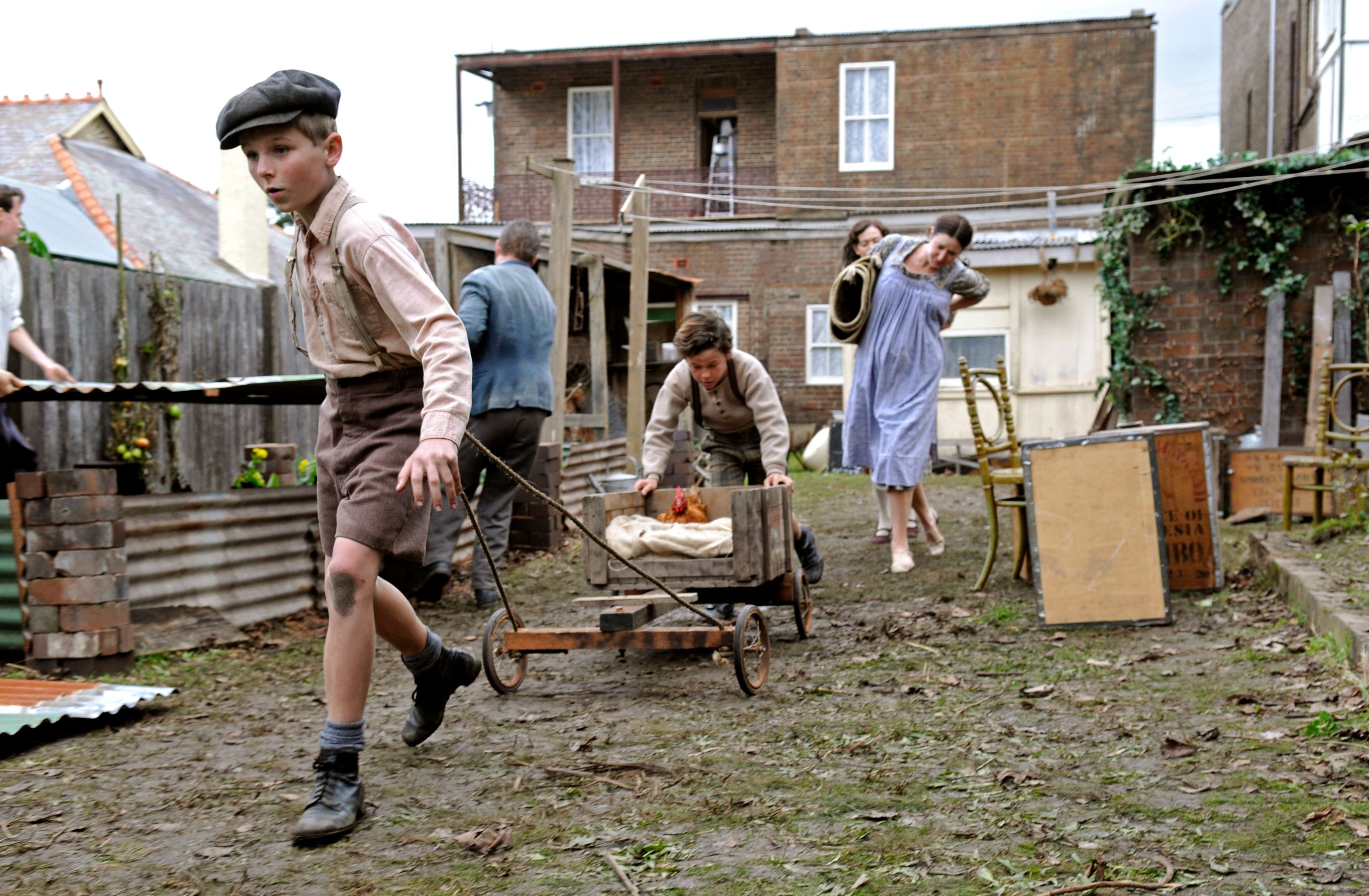 A group of children and adults are engaged in various activities in a muddy outdoor area filled with makeshift structures. One boy runs away, wearing a cap, while another pulls a cart with a chicken inside. Nearby, a young girl in a blue dress walks with a seated person, and an adult appears to be carrying a stringed instrument. The setting conveys a bustling, informal atmosphere.