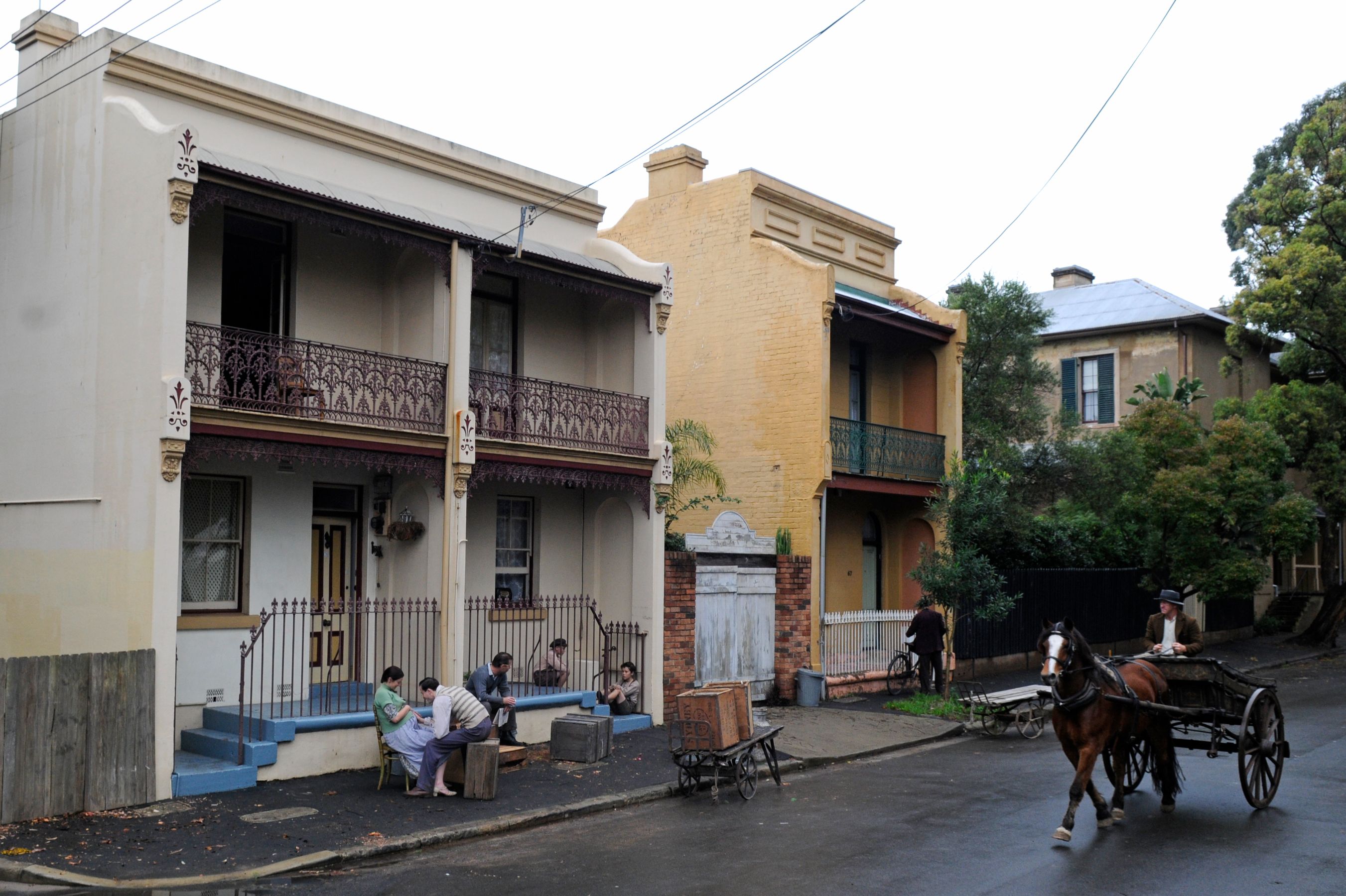 A vintage street scene features two two-story buildings with decorative balconies and an inviting entrance. Several people, dressed in period attire, sit on the steps, engaged in conversation, while a horse-drawn cart moves along the quiet street, enhancing the nostalgic atmosphere. Lush greenery and other nearby structures create a charming backdrop.