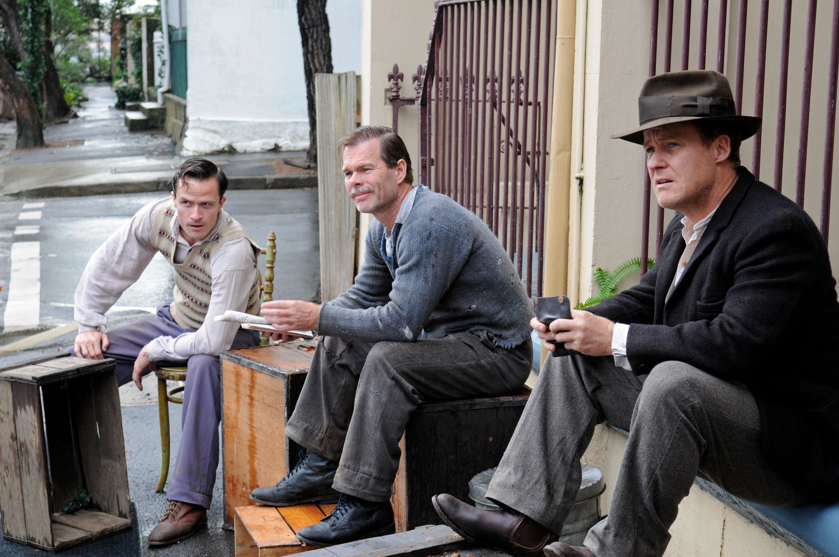 Three men sit on wooden crates outside a building, appearing contemplative. One man wears a sweater and holds a piece of paper, while another dressed in a suit and fedora looks thoughtfully into the distance. The scene is set on a narrow street lined with trees.