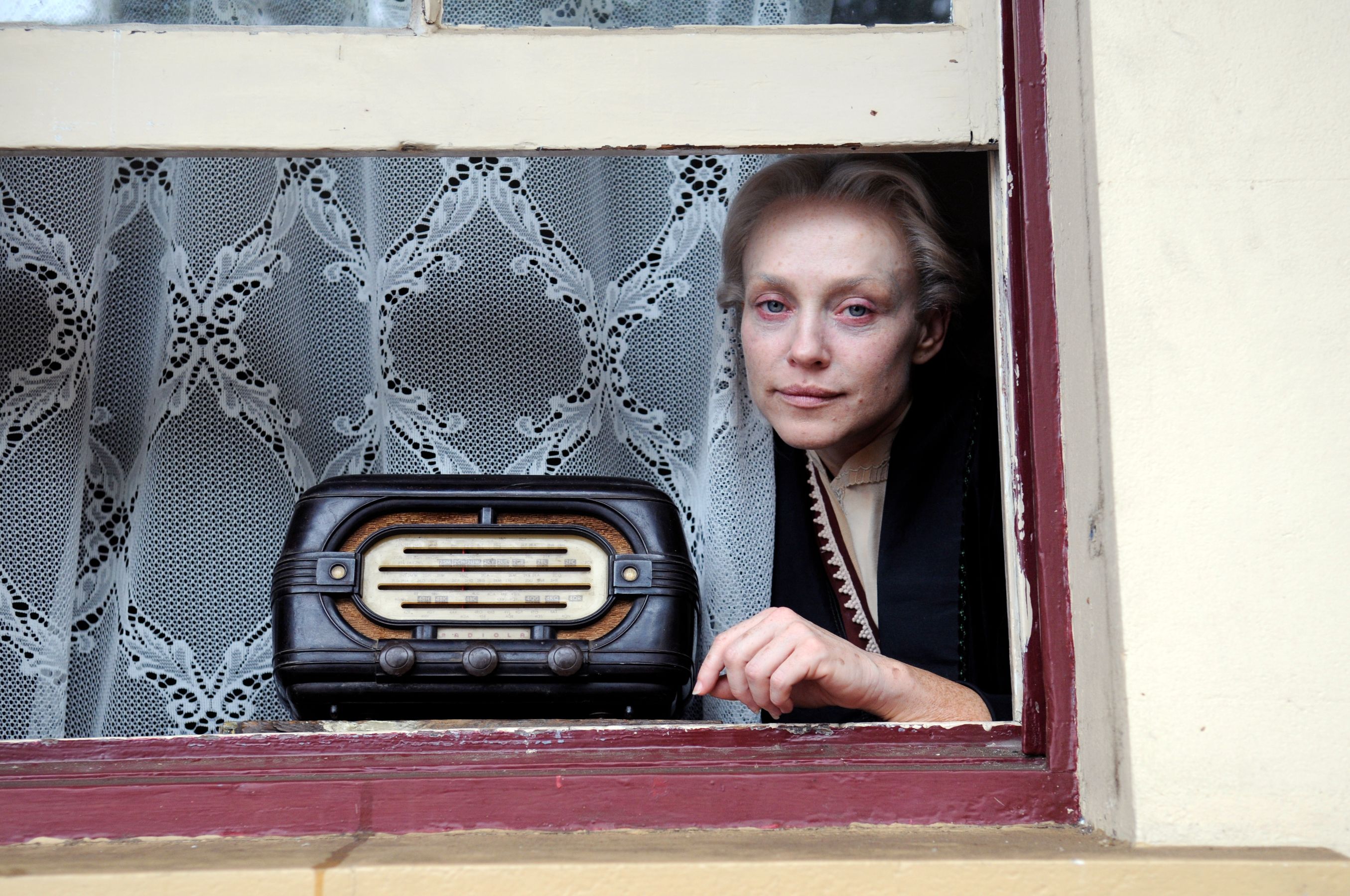 A woman with pale skin and light blonde hair gazes out from a window adorned with lace curtains. She rests her hand on the ledge near an old-fashioned radio that sits nearby.