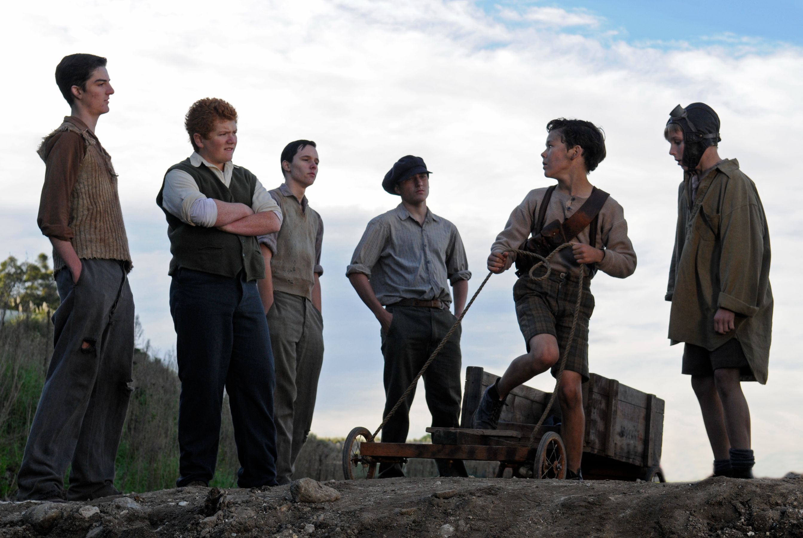 A group of six boys, dressed in early 20th-century clothing, stand in an outdoor setting. One boy is actively engaged with a wooden cart, while the others observe him with various expressions, suggesting a moment of camaraderie or play. The background features a cloudy sky and sparse vegetation.