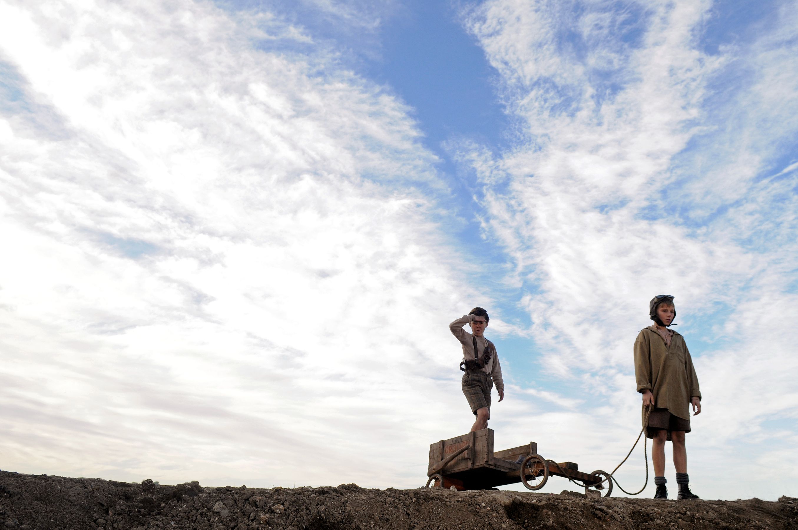 Two boys stand on a raised dirt surface under a cloudy sky. One boy looks out as if scouting the area while the other stands nearby, holding a rope attached to a small wooden cart.