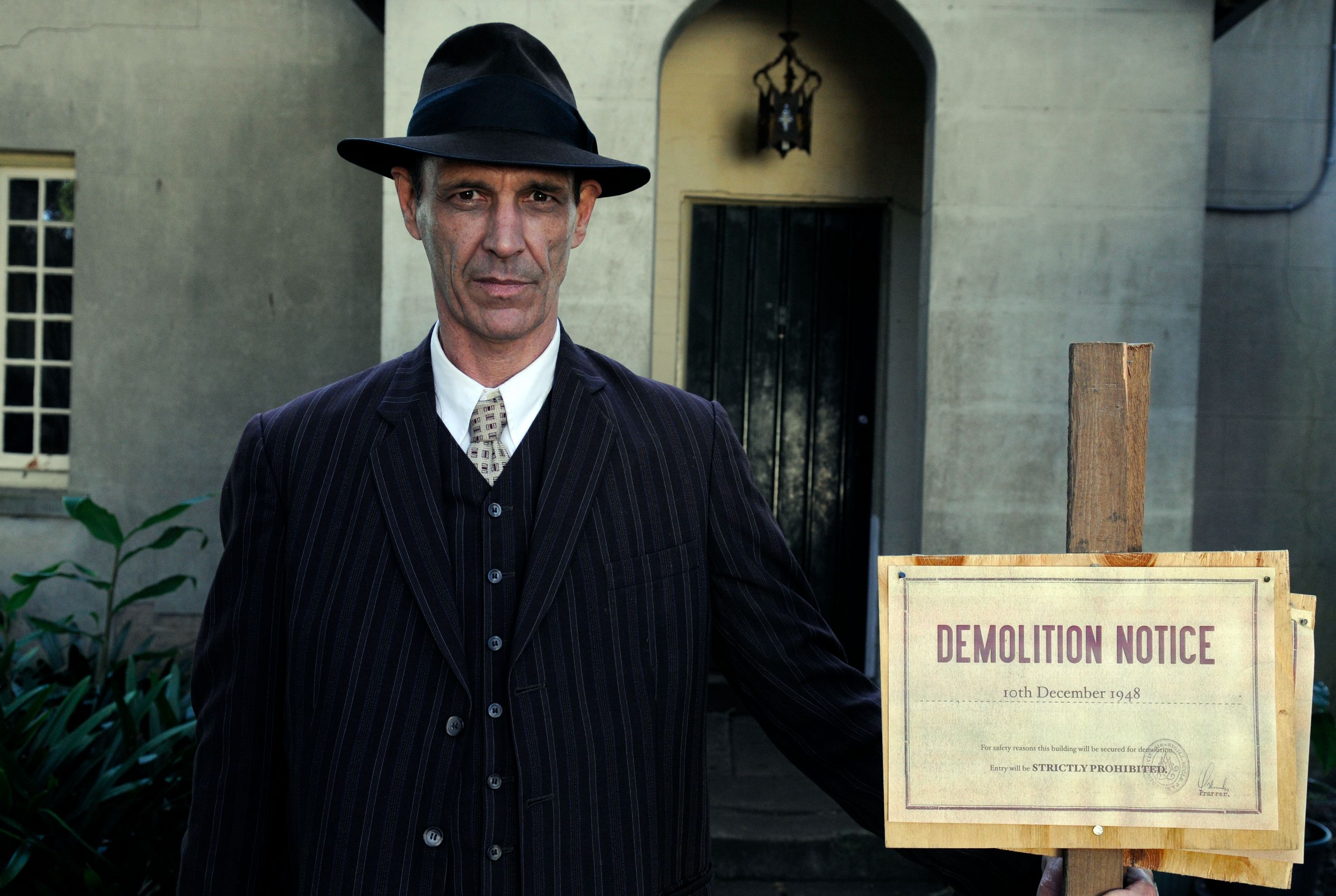 A man in a black pinstriped suit and a fedora stands in front of a building, holding a wooden sign that reads "DEMOLITION NOTICE" with a date of December 10, 1948. The background features a dark door and light-colored walls.