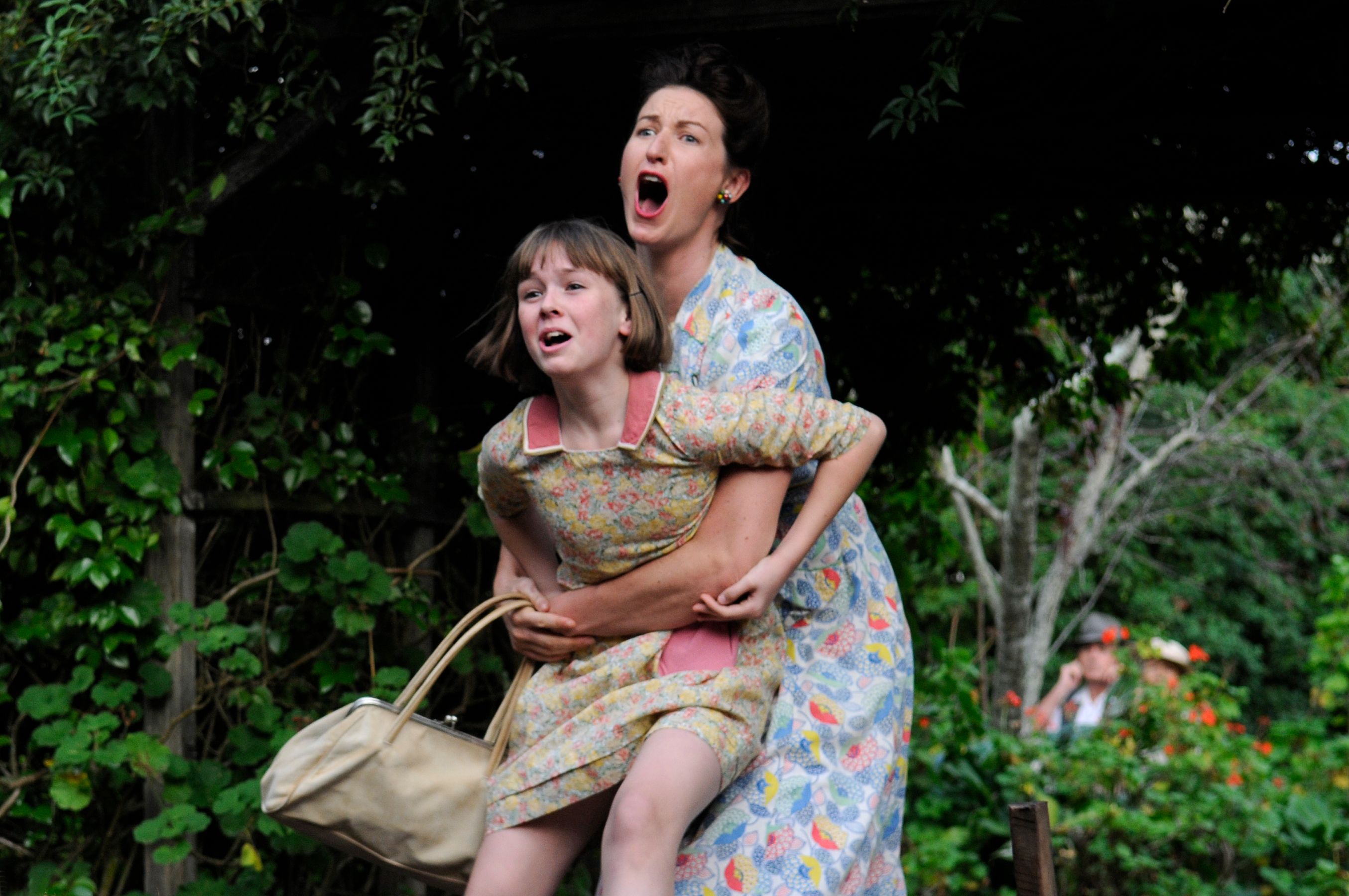 A woman wearing a patterned dress joyfully embraces a young girl in a similar patterned outfit, both laughing loudly. The background is filled with lush greenery, suggesting a warm, outdoor setting.