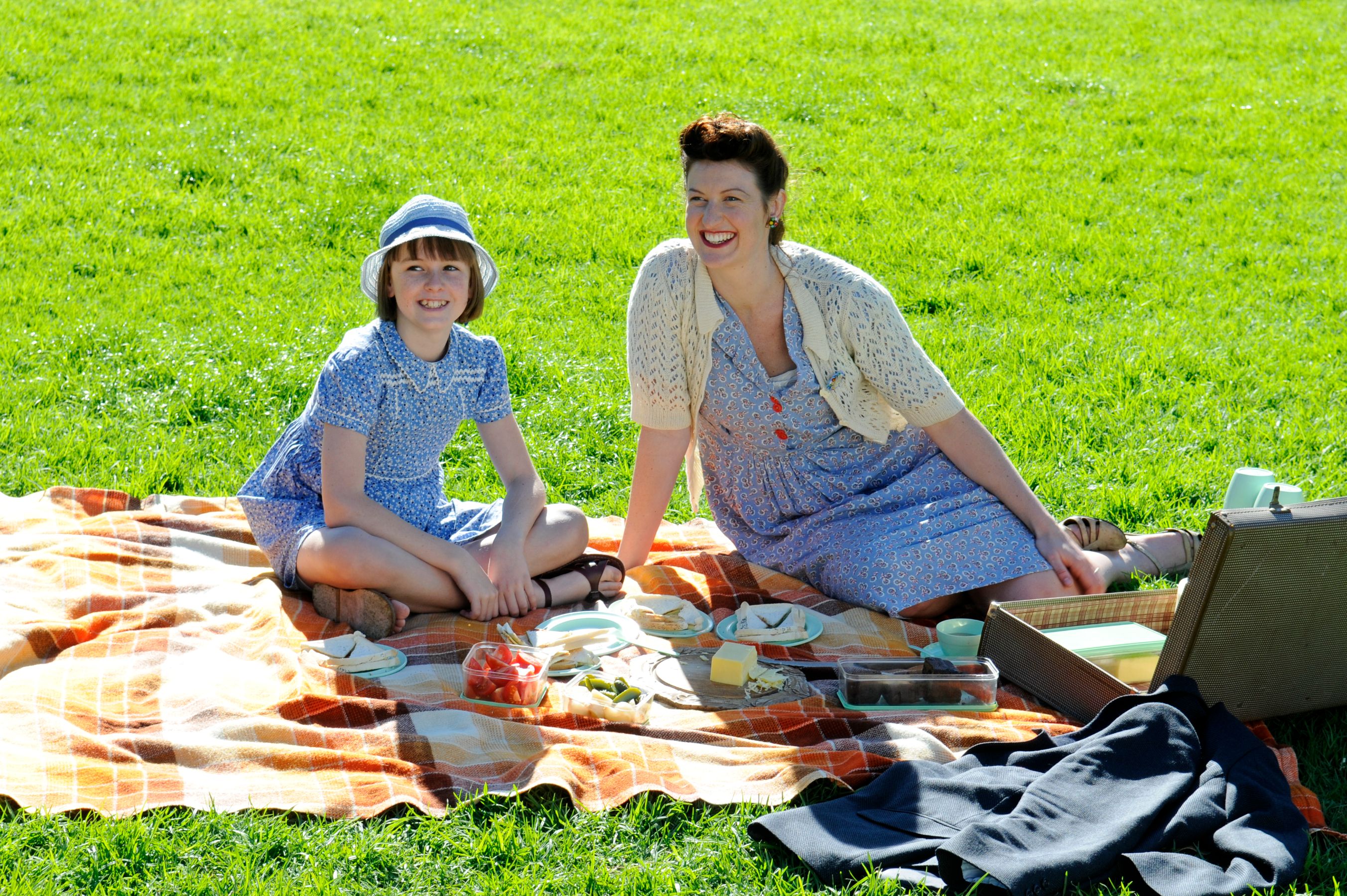 A woman and a girl sit on a picnic blanket in a sunny park, smiling at the camera. They are surrounded by an assortment of food, including cheese, fruit, and sandwiches, with a picnic basket nearby. The grassy background enhances the cheerful atmosphere.