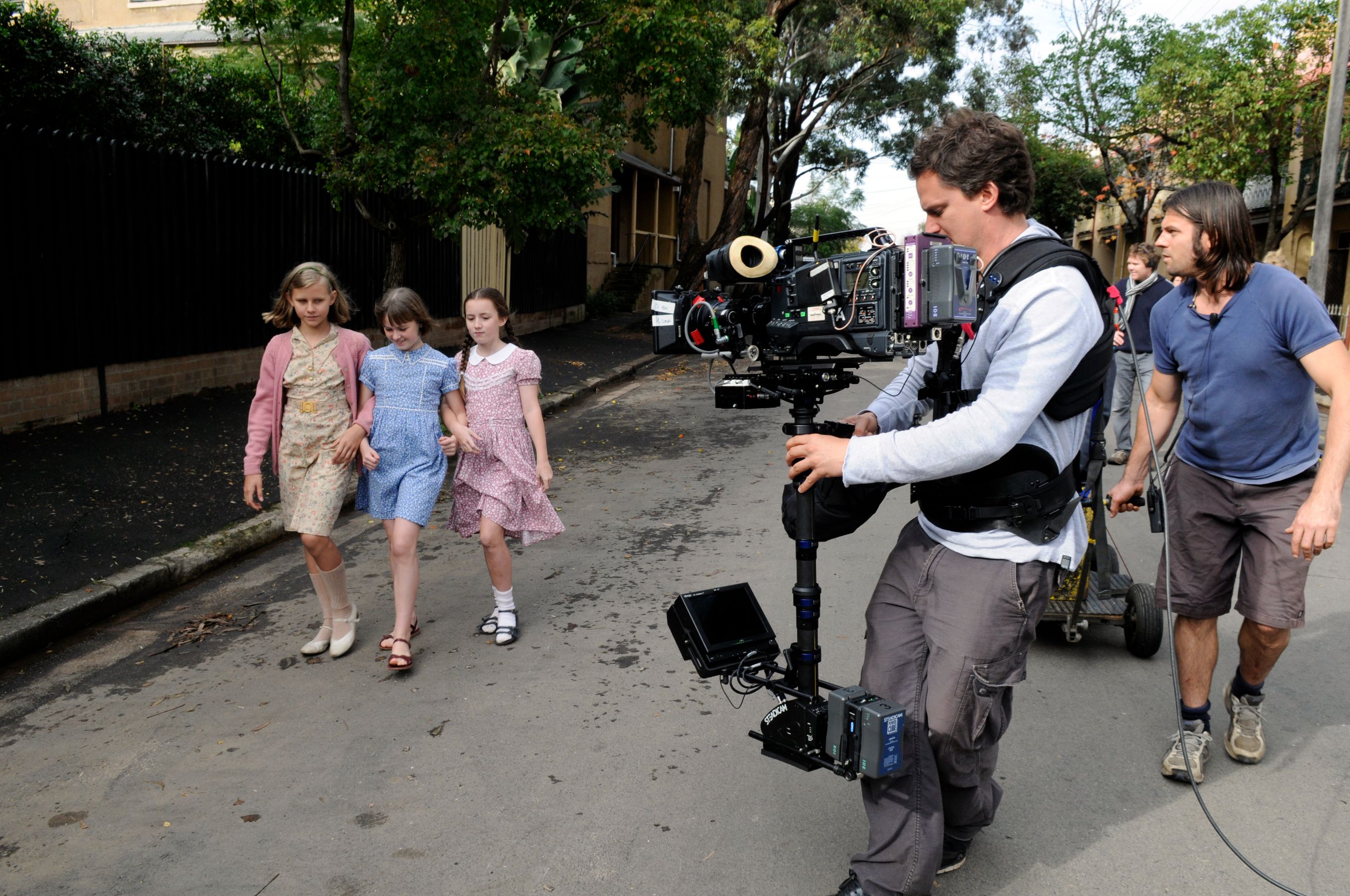 Three children dressed in vintage-style dresses walk together down a street. A cameraman equipped with a professional camera rig follows them closely, while another crew member assists in the background. Trees and buildings line the street, creating a lively filming atmosphere.