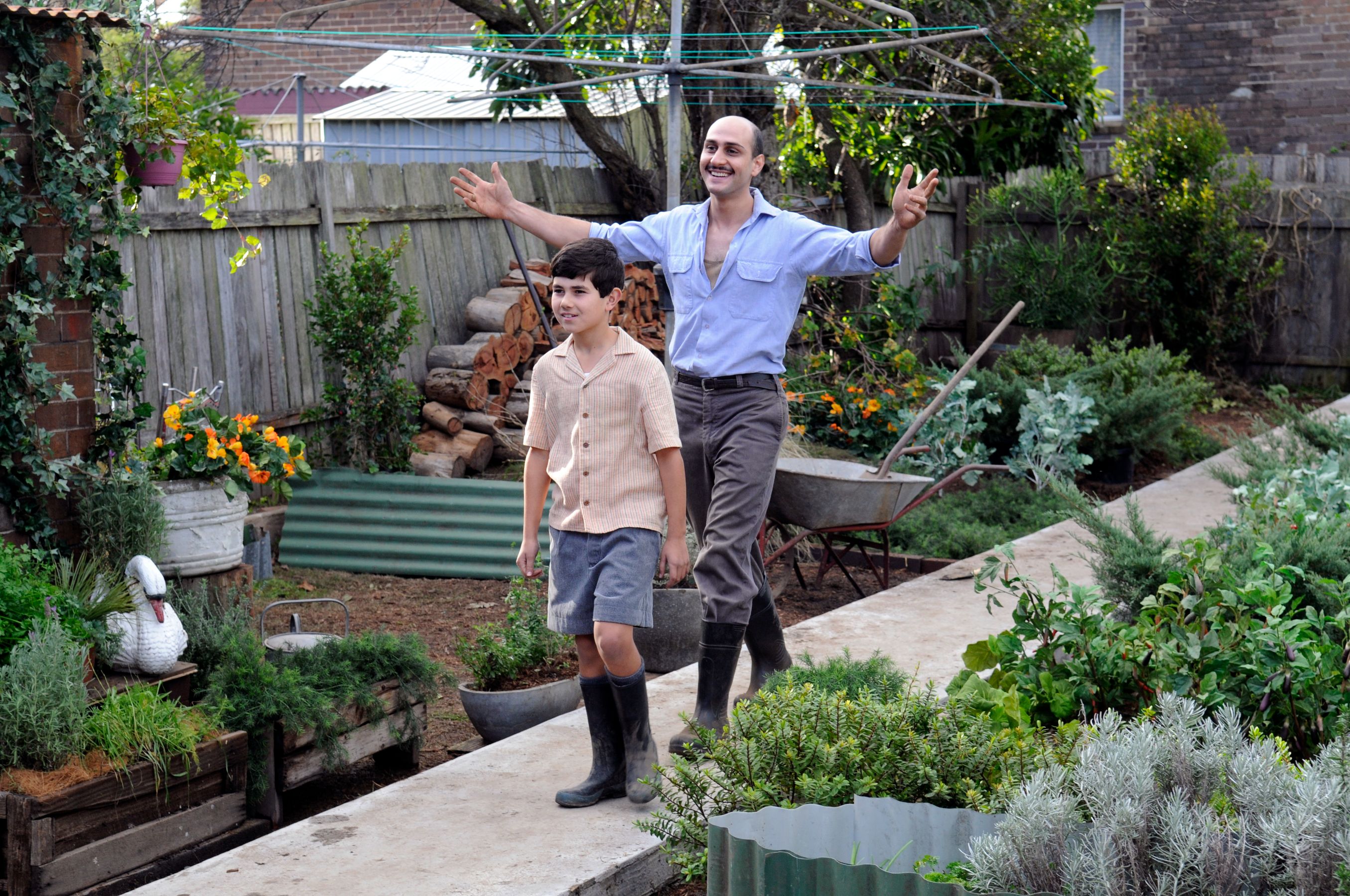 A joyful adult man and a young boy walk along a stone path through a lush garden filled with various plants and flowers. The man has his arms outstretched, expressing enthusiasm, while the boy walks beside him with a smile, both dressed casually.