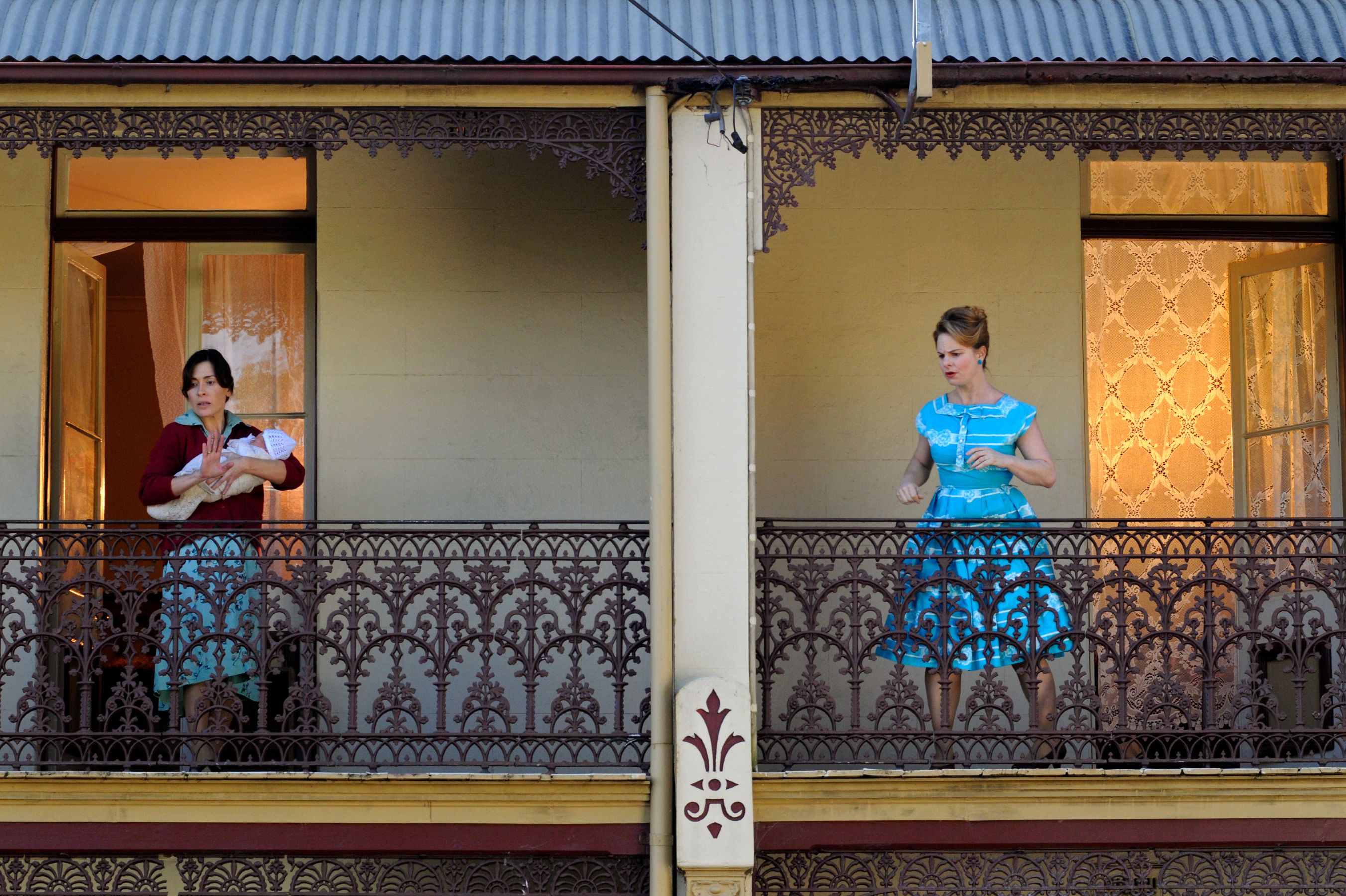 Two women stand on their respective balconies of a decorative Victorian-style building. On the left, one woman cradles a baby while wearing a red sweater and skirt, looking concerned. The other, on the right, is dressed in a blue dress with white stripes and appears to be in motion, seemingly engaged in conversation or thought. The warm glow from an interior window contrasts with the intricate metalwork of the balconies.