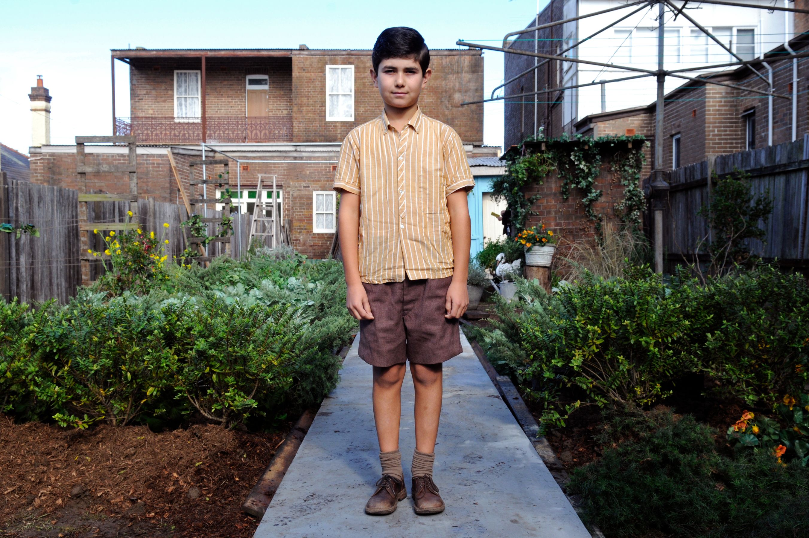 A young boy stands on a concrete pathway in a garden, wearing a striped yellow shirt, brown shorts, and brown shoes. Lush greenery is on either side of the path, with a backdrop of brick houses and wooden fences.
