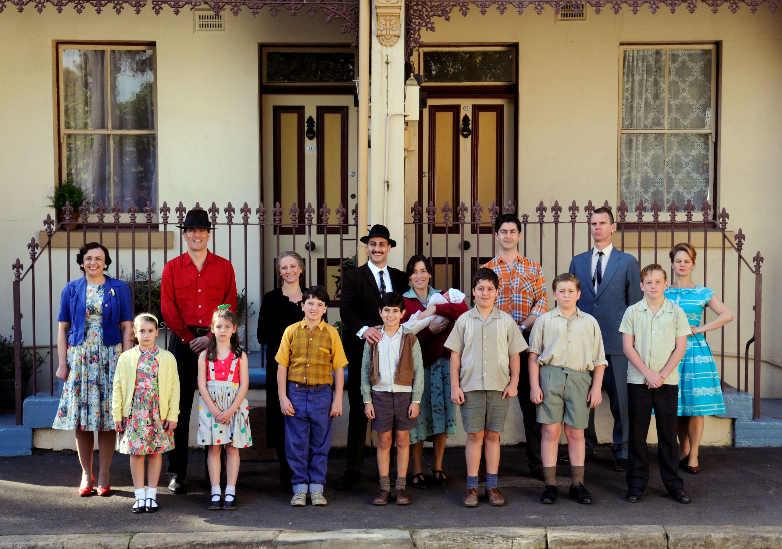 A group of adults and children stands together in front of a vintage-style building with a wrought iron fence. The scene features individuals in colorful, retro clothing, with some children holding hands and others posing confidently. The group appears to be part of a production or family gathering, with expressions ranging from cheerful to serious.