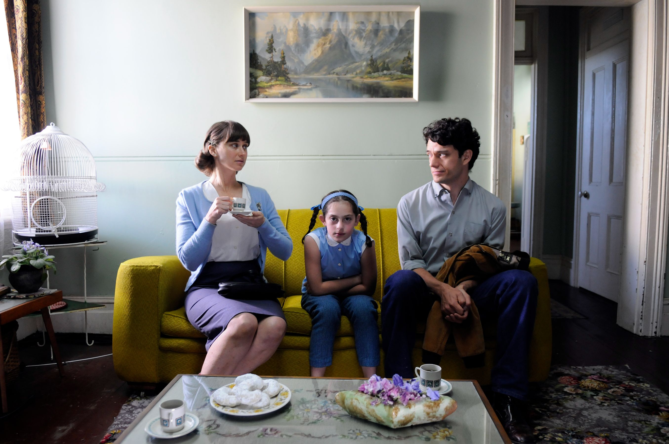 A woman in a light blue cardigan holds a cup while sitting on a bright yellow couch, looking at a man beside her. A girl in a blue dress sits between them, appearing pensive with her arms crossed. The setting is a cozy living room featuring a birdcage, a small table with pastries, and a landscape painting on the wall.