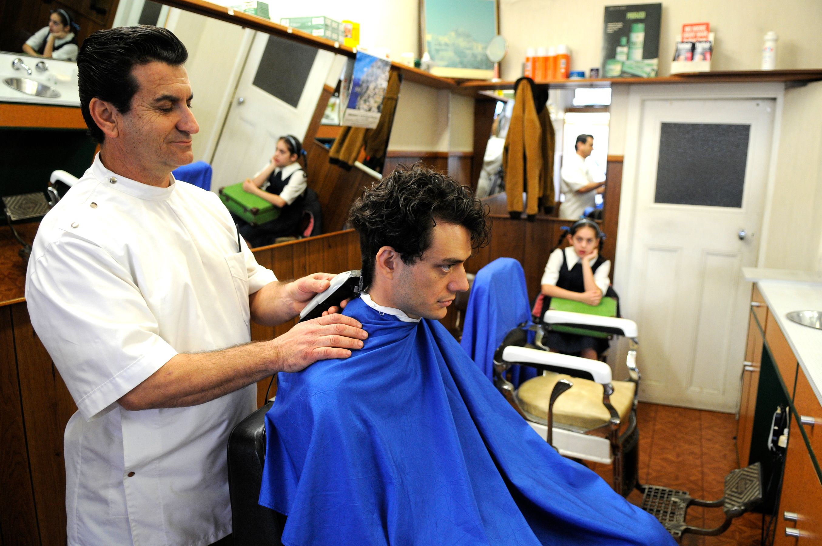 A barber in a white coat is using clippers on the hair of a young man draped in a blue cape. In the background, two young girls sit in chairs, one holding a green backpack, while the other looks on attentively. The barbershop has wooden paneling and mirrors reflecting the bustling environment.