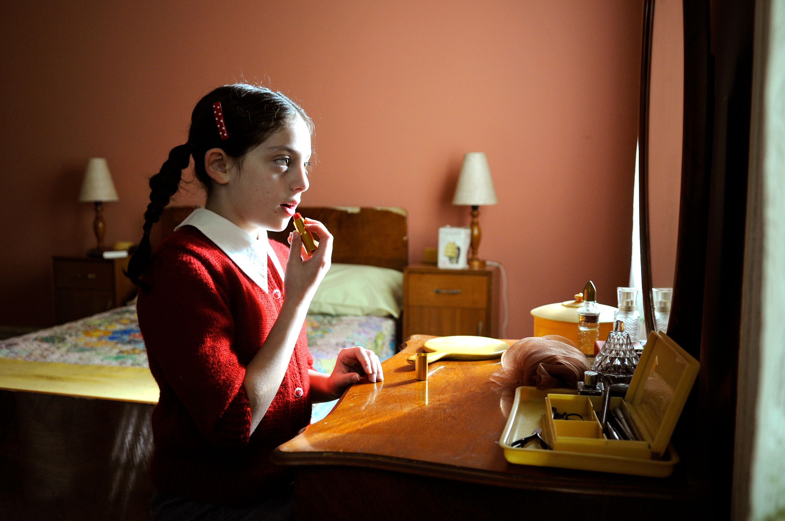 A young girl with braided hair and wearing a red cardigan applies lipstick while looking into a mirror. The background features a softly lit bedroom with a bed covered in a colorful quilt, bedside lamps, and various personal items on the dresser, creating a nostalgic atmosphere.
