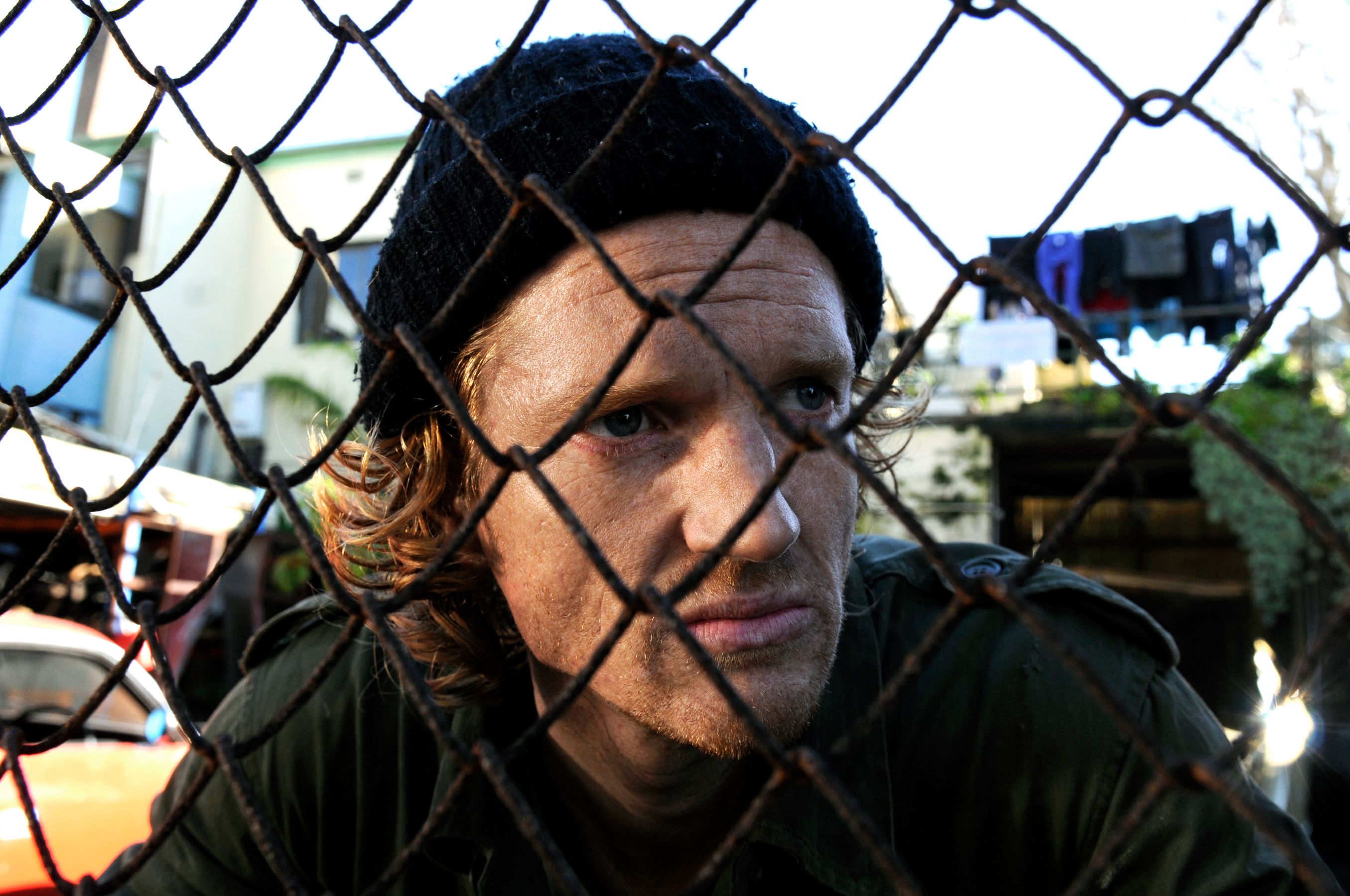 A man with curly hair and a black beanie gazes thoughtfully through a rusted wire fence, his expression serious. In the background, blurred hints of colorful clothing and structures suggest a gritty urban environment.