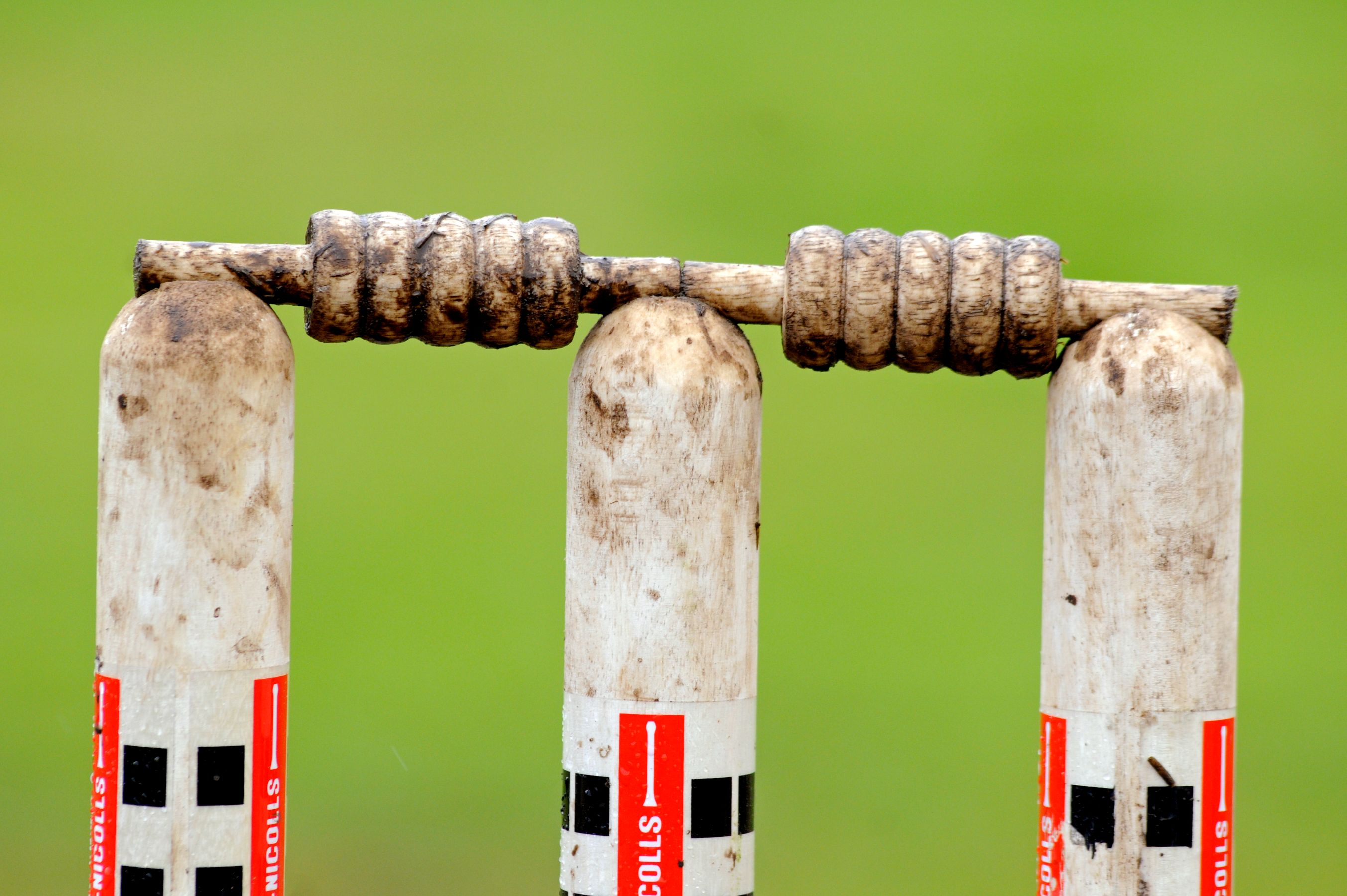 Three white cricket stumps stand upright, topped with a wooden bails that are wrapped with cord. The stumps show signs of wear, with dirt smudges visible on their surfaces, and the background is a soft, blurry green.