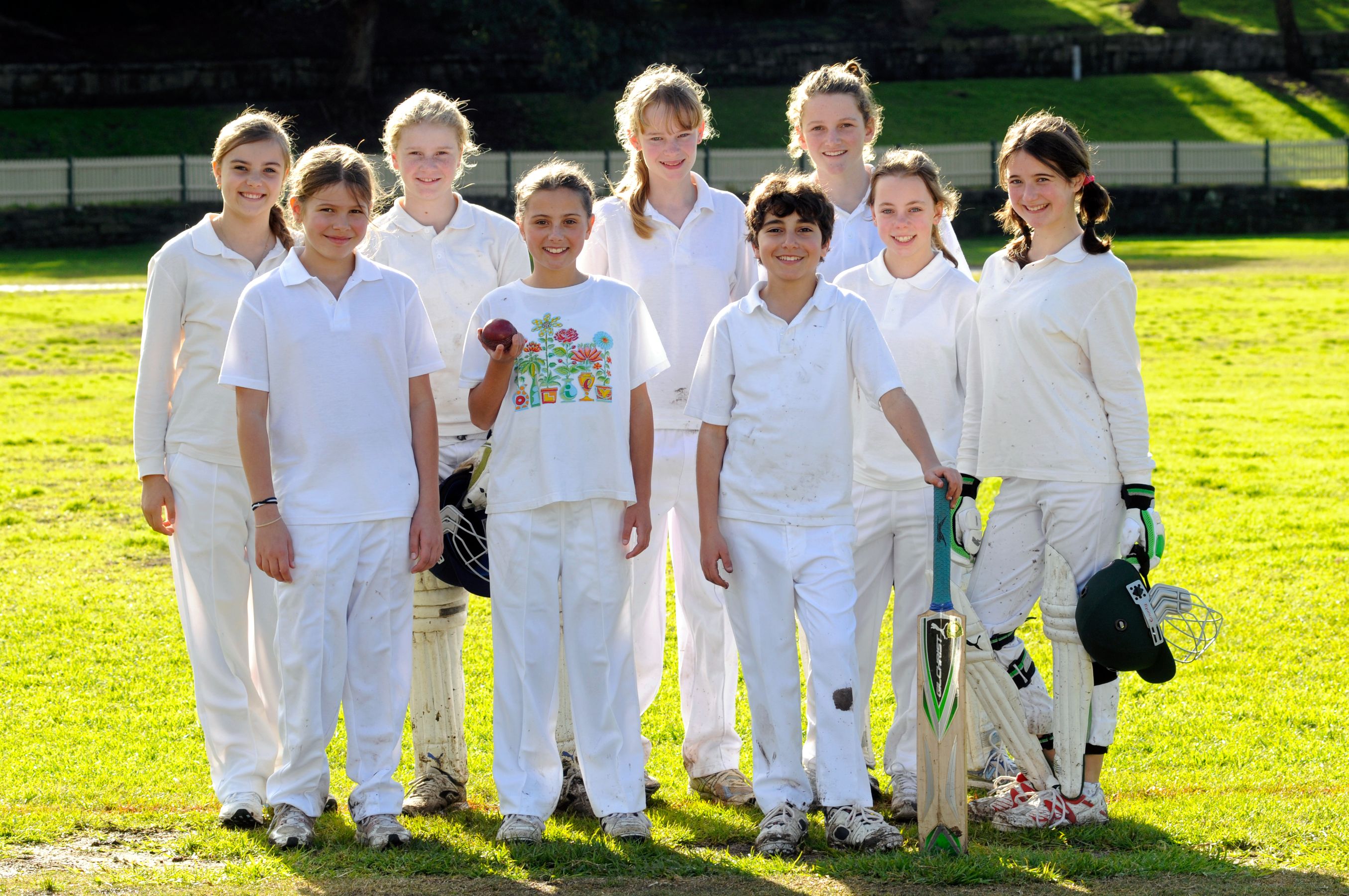 A group of ten young cricketers, dressed in white cricket uniforms, pose together on a grassy field. One girl holds a cricket ball, while another has a bat. They smile at the camera, showcasing camaraderie and youthful enthusiasm for the sport.