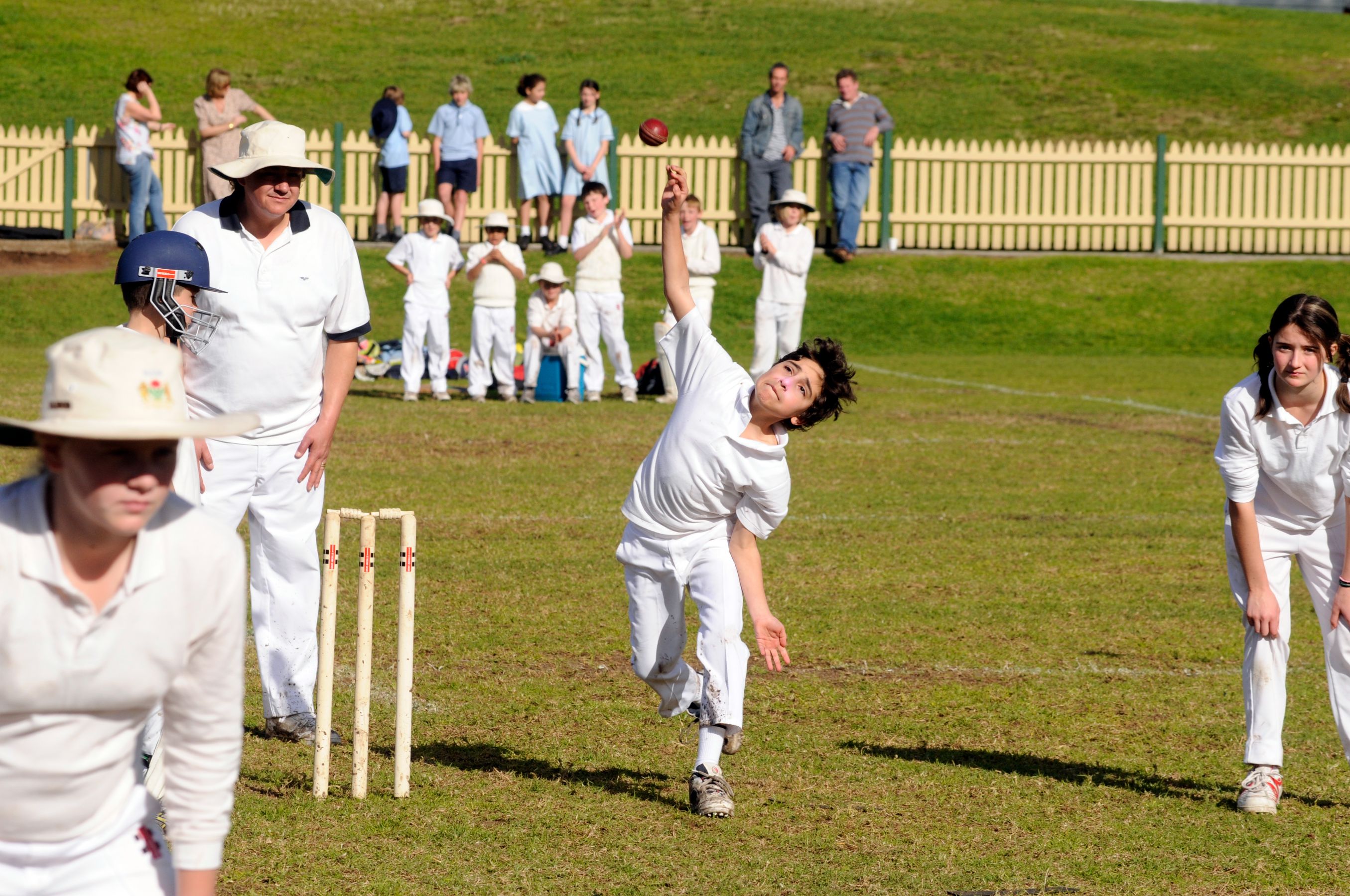 A young cricketer bowls a ball on a grassy field, wearing a white uniform and a hat. Nearby, a boy in a batting helmet prepares to receive the delivery, while the bowler's teammates watch from various positions around the field, with spectators in the background.