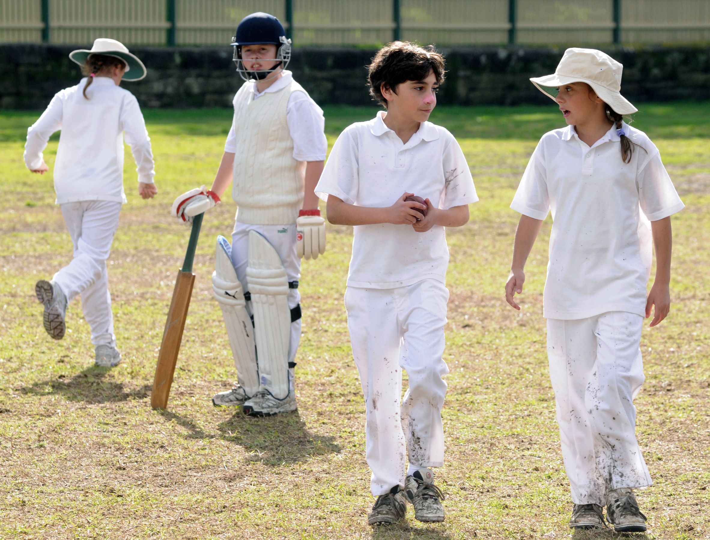 A group of children in white cricket uniforms engage in a casual conversation on a grassy field. One child, wearing a cricket helmet and pads, stands with a bat, while two others walk nearby, one of them sporting a wide-brimmed hat. Another child, having turned away, walks toward the boundary of the field. The scene captures the playful and relaxed atmosphere of youth sports.