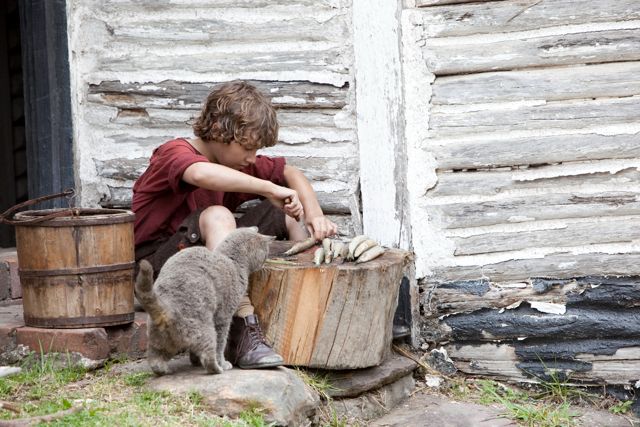 A young boy dressed in a red shirt sits on a wooden stump, focused on carving a piece of wood. A gray cat approaches him, curiously watching the activity. In the background, rustic wooden walls and two barrels add to the rural setting.