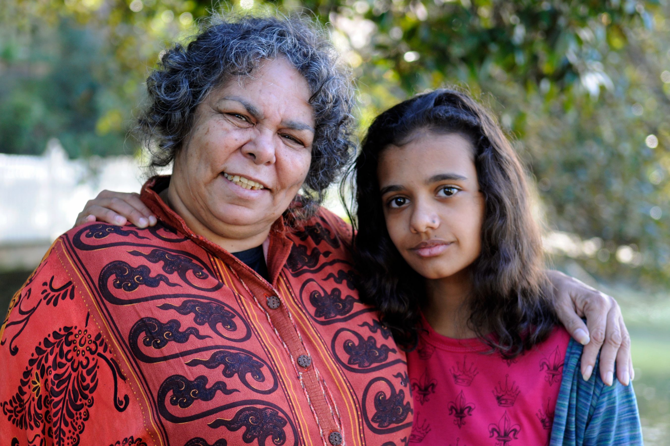 A woman with curly hair wearing a brightly patterned red garment smiles while standing next to a girl with long, dark hair, who wears a pink shirt and looks at the camera. The two appear close, with the woman resting her hand on the girl's shoulder, set against a backdrop of greenery.