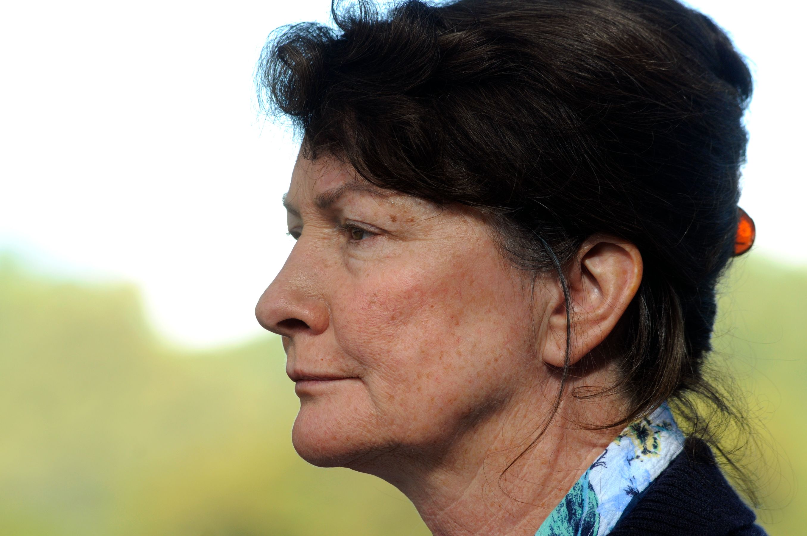 A woman with dark, shoulder-length hair is shown in profile, wearing a patterned scarf underneath a blue sweater. She has a neutral expression, with visible freckles and an ear adorned with a small orange clip. A soft, blurred background adds to the tranquil composition.