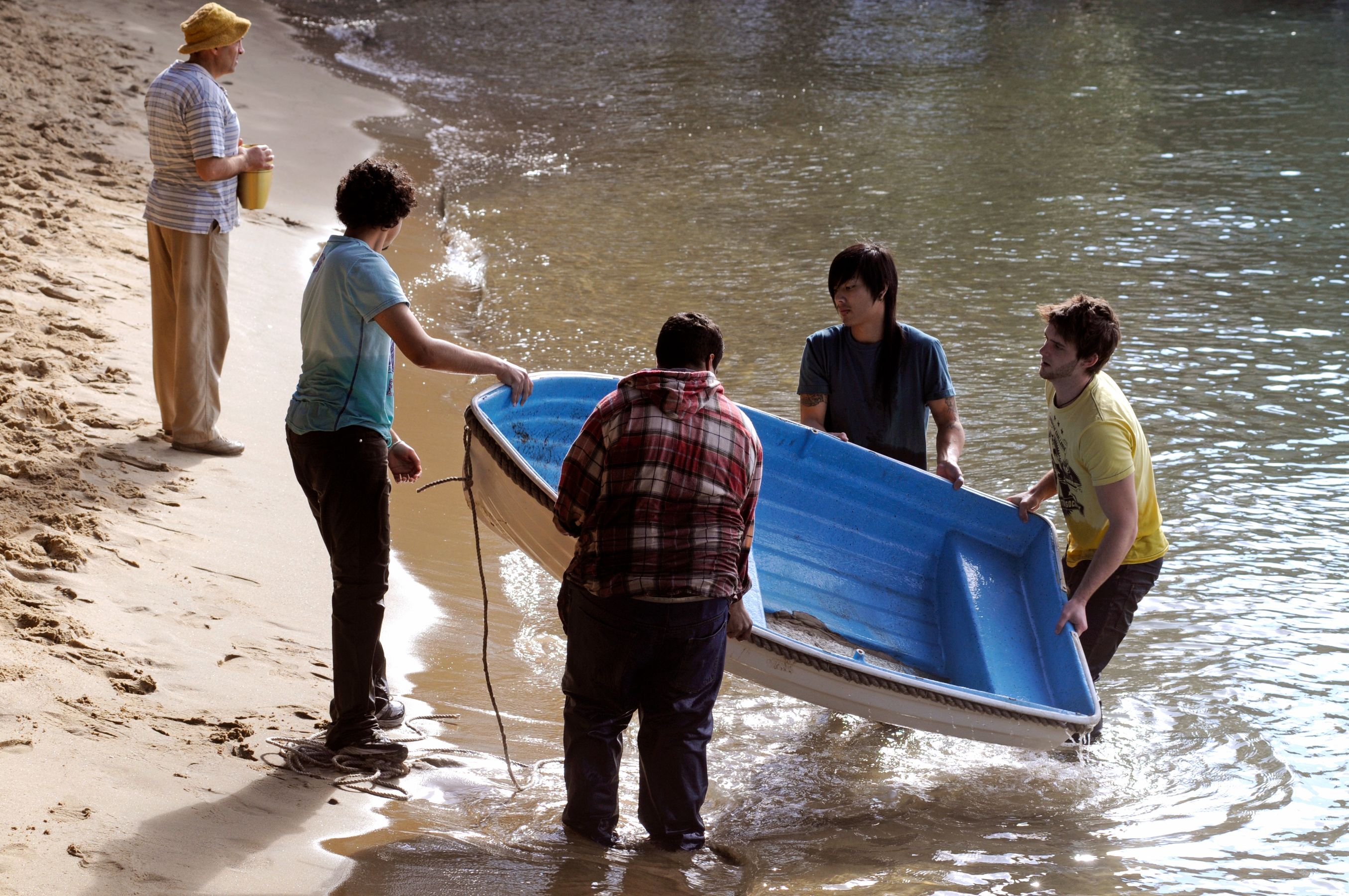 Four individuals are lifting a small blue boat from the water onto the sandy shore, while a fifth person stands nearby observing. The scene is set by a calm body of water, with gentle waves reflecting sunlight, and the group appears to be working together.