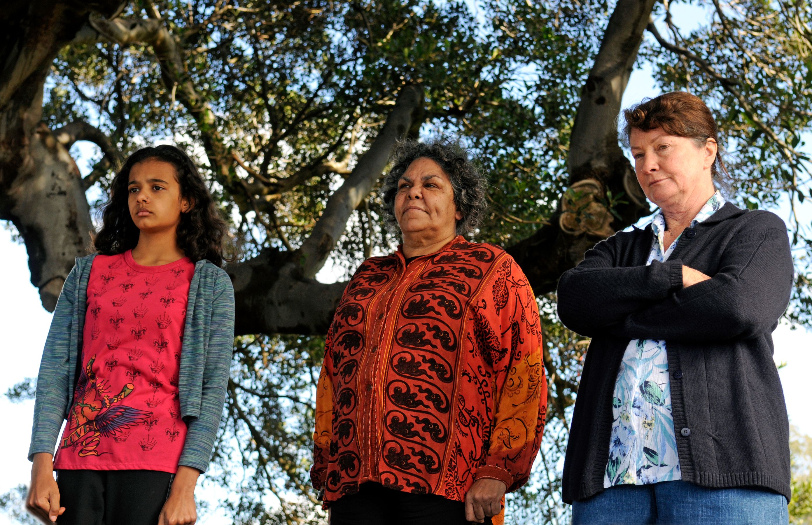 Three women stand together outdoors under a tree, each displaying a serious expression. The woman on the left, a girl with curly hair, wears a bright red shirt with a design. The woman in the middle, an adult with curly hair, is dressed in an intricately patterned orange blouse, while the woman on the right, an older adult with straight hair, is wearing a dark cardigan over a floral blouse. The background features leafy branches framing the scene.