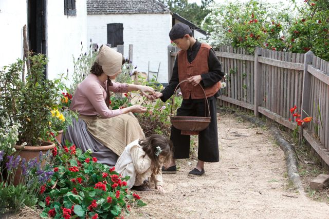 A woman in historical attire interacts with a boy, who is holding a woven basket, while a young girl plays in the dirt nearby. Brightly colored flowers line the dirt path, and a white cottage with a thatched roof is visible in the background.