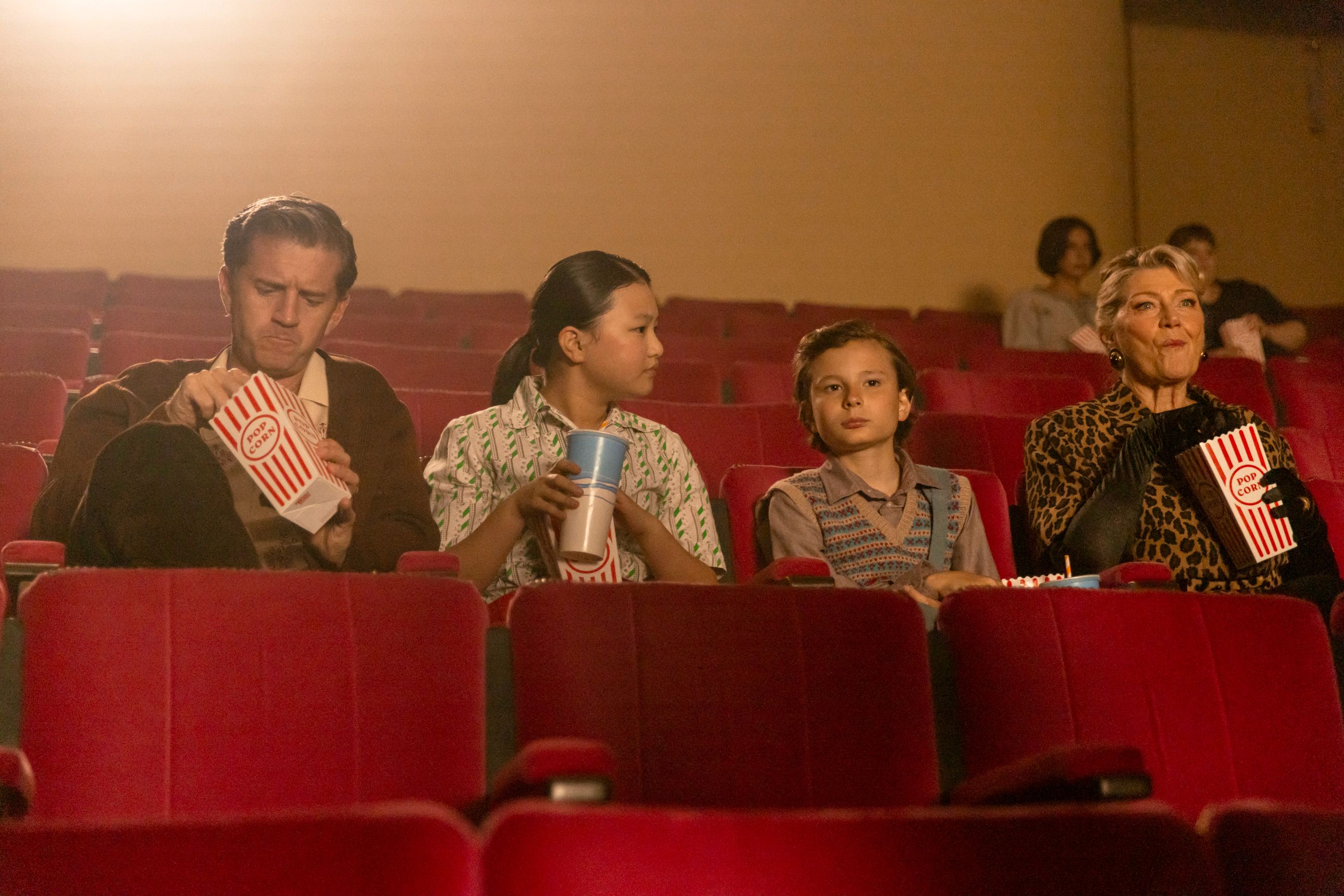 A group of four people sits in a nearly empty movie theater, each holding snacks. The two children are in the middle, with a boy looking attentively and a girl holding a drink. An older man on the left is focused on a popcorn box, while an older woman on the right enjoys her popcorn, dressed in a stylish coat. Soft lighting enhances the nostalgic atmosphere of the scene.
