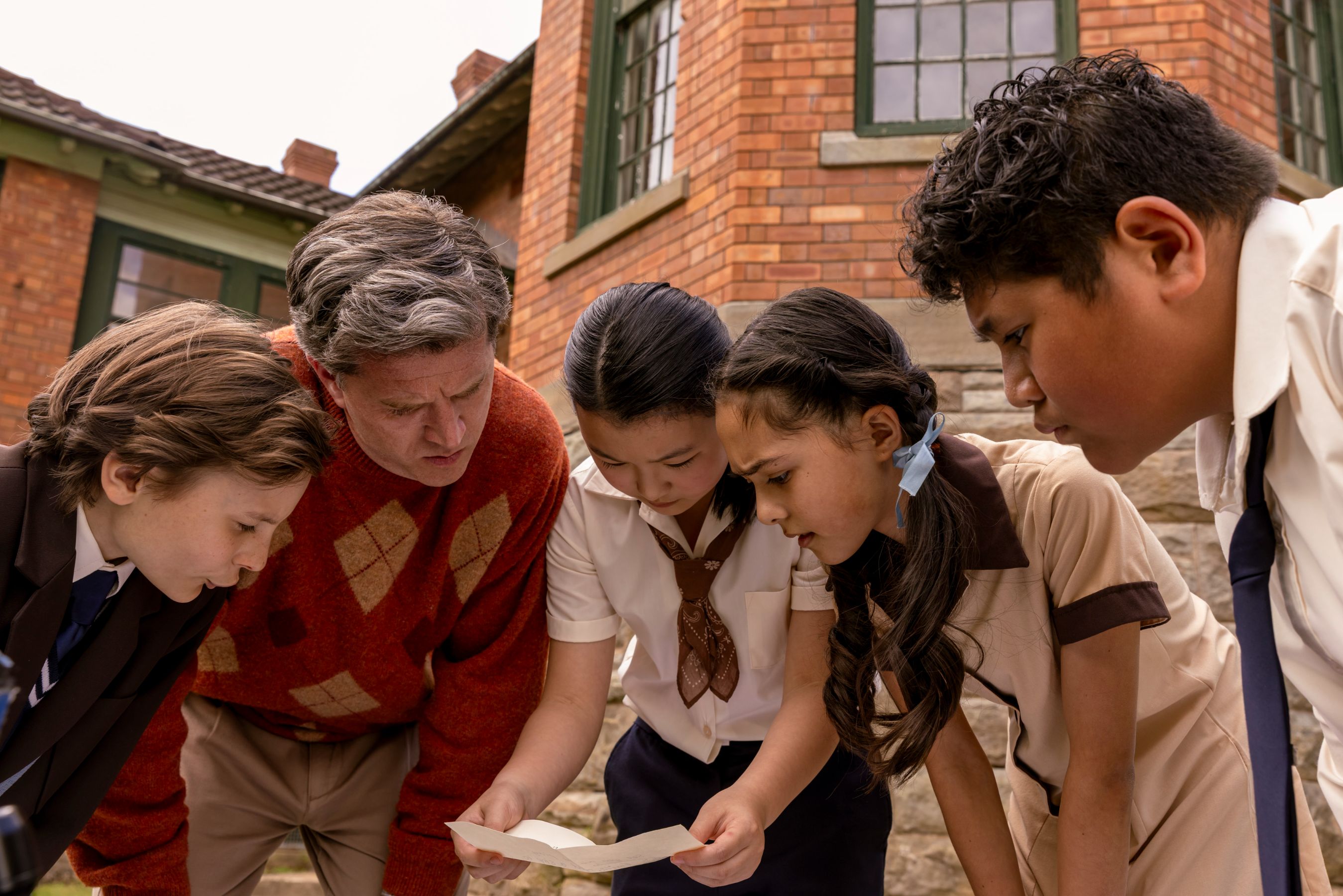A group of four children and one adult huddle closely together, examining a piece of paper intently. They are outdoors near a brick building, with expressions of curiosity and focus as they engage with the document.