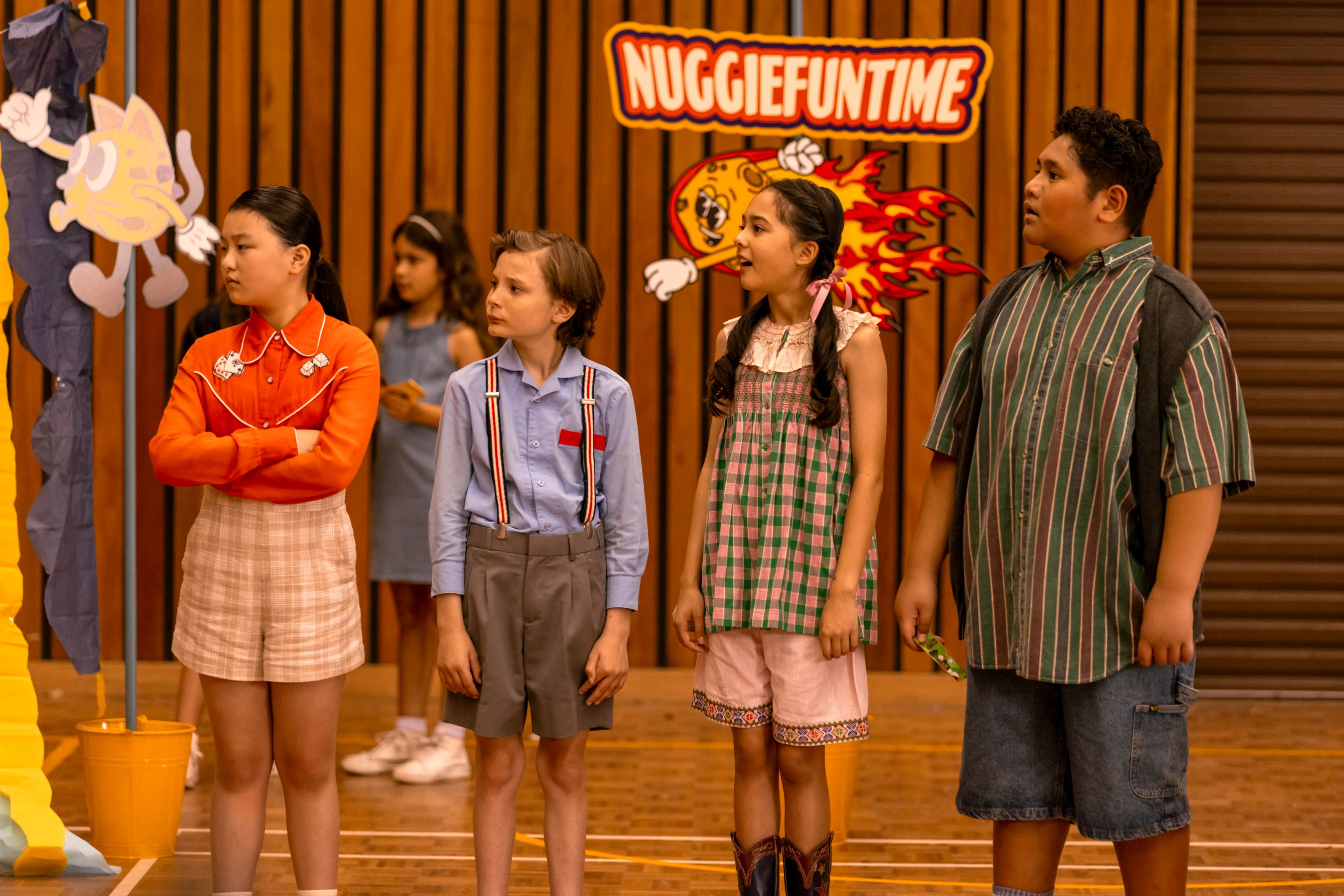 A group of five children stands together in a gymnasium, looking towards something off-screen. The girl in a red top and plaid shorts has her arms crossed, while another boy wears suspenders and looks curious. A girl in a striped dress appears surprised, and the boy next to her, wearing a striped shirt, seems intrigued. In the background, a colorful banner reads "Nuggiefuntime." The walls are made of wooden panels, and other children are visible in the background.