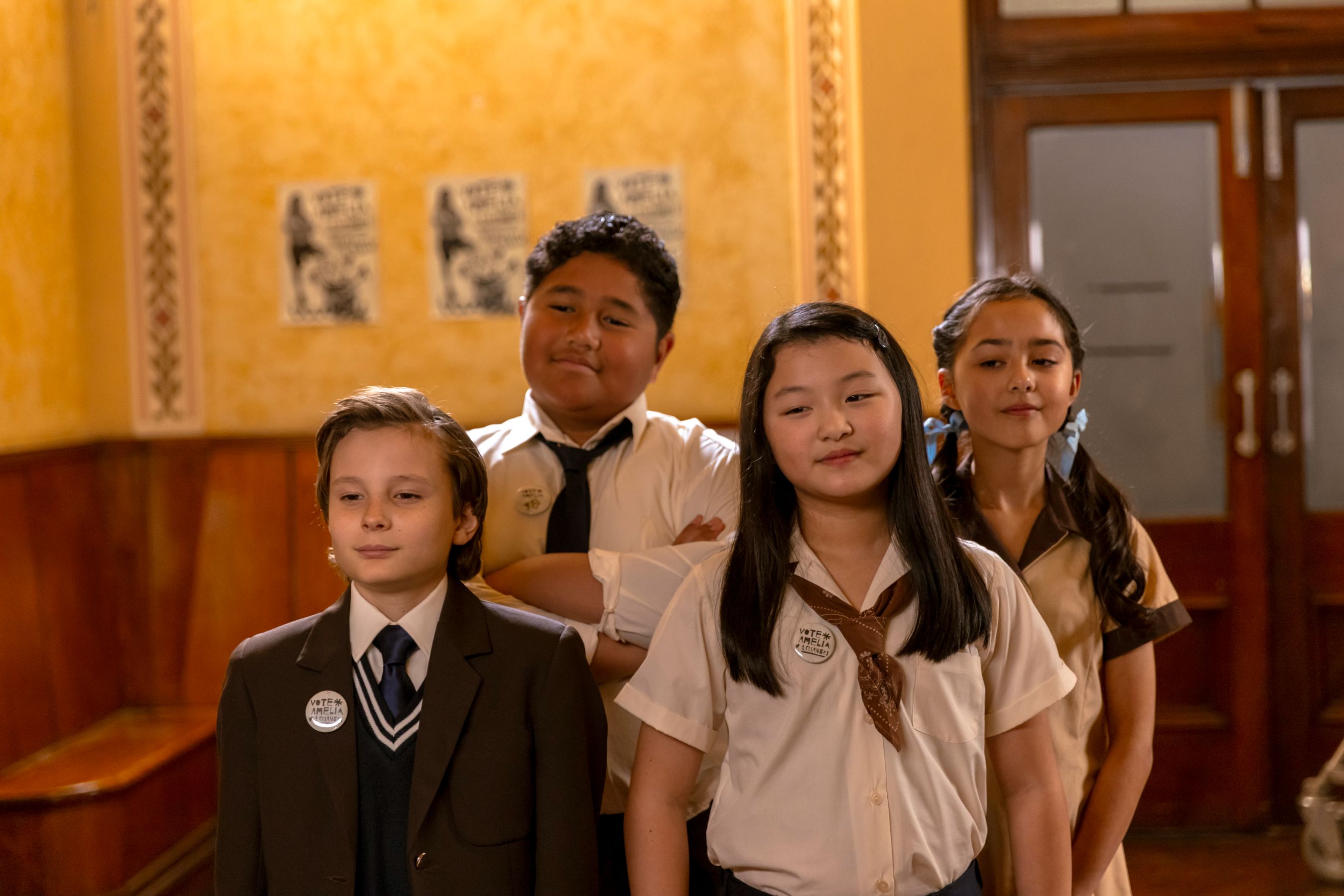 Four children stand together in a warmly lit hallway with yellow walls. They wear school uniforms and badges, giving off a confident demeanor as they pose for the photo. Posters can be seen in the background, adding to the school atmosphere.