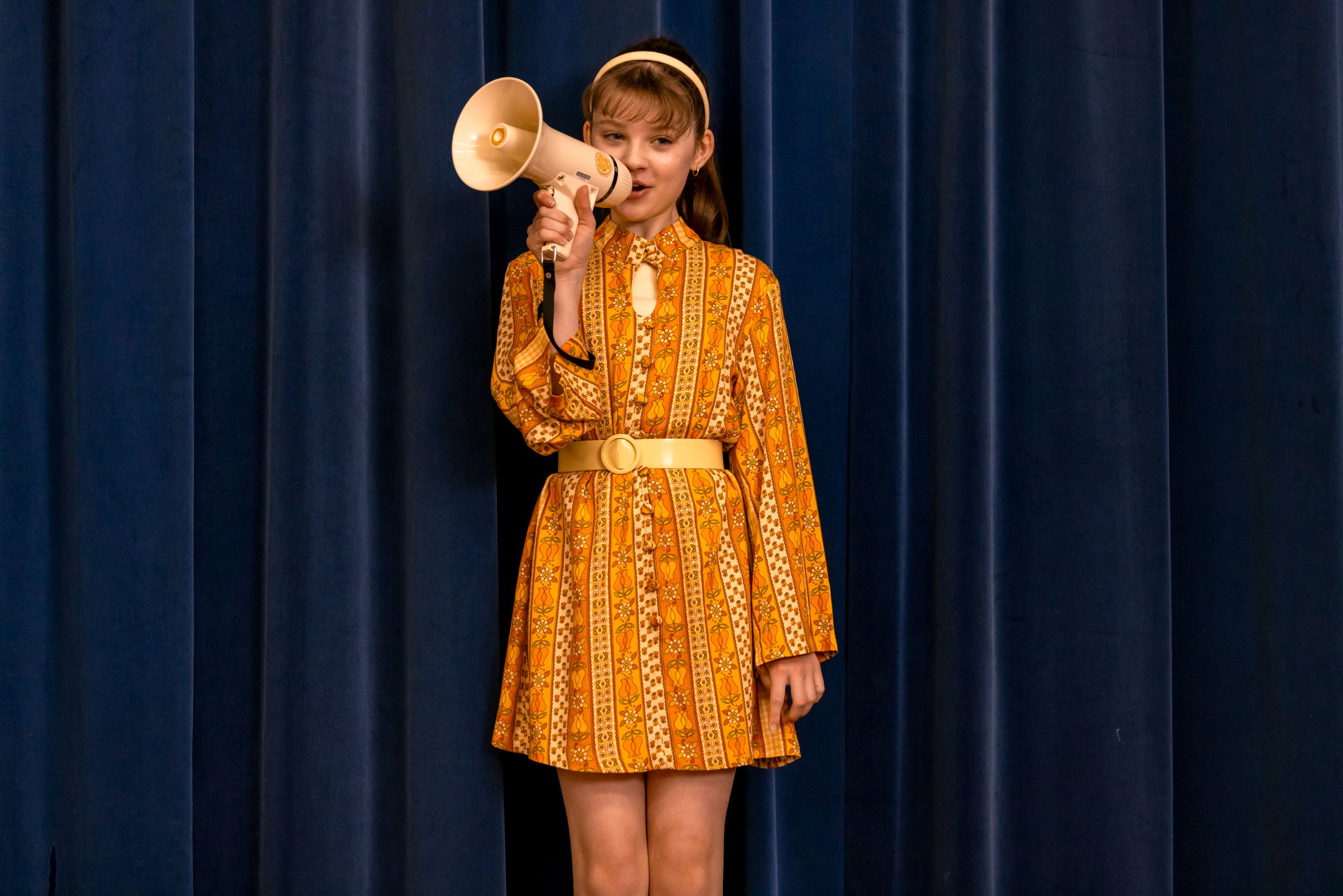 A young girl stands on a stage in front of dark blue curtains, wearing a vintage-style orange dress with floral patterns and a yellow belt. She holds a megaphone to her mouth, appearing to address an audience.