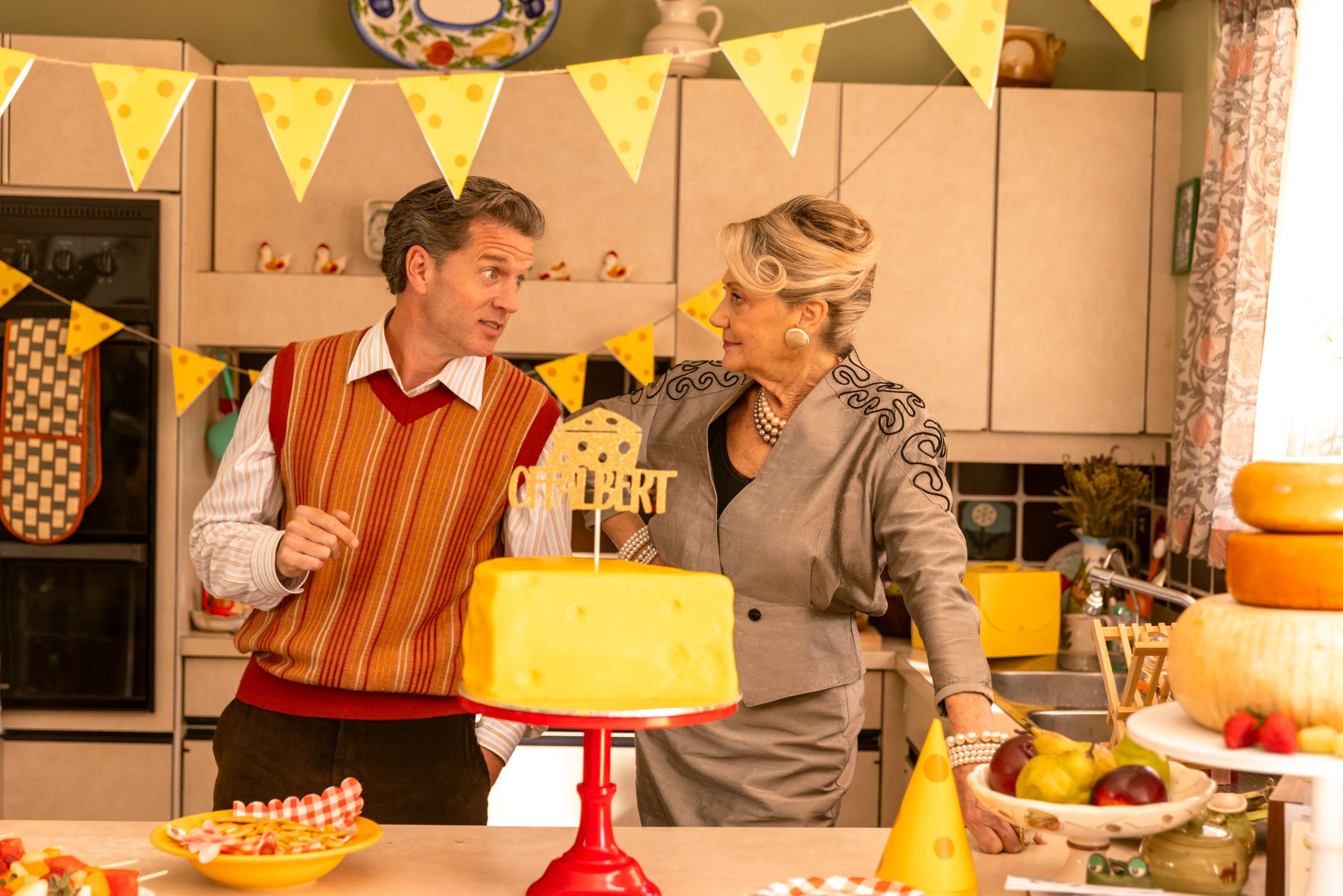 A man in a striped vest and a woman in a gray outfit with elaborate detailing engage in conversation in a brightly decorated kitchen. A large yellow cake resembling cheese sits on a stand between them, surrounded by festive yellow bunting. Various fruits and snacks are displayed on the counter.