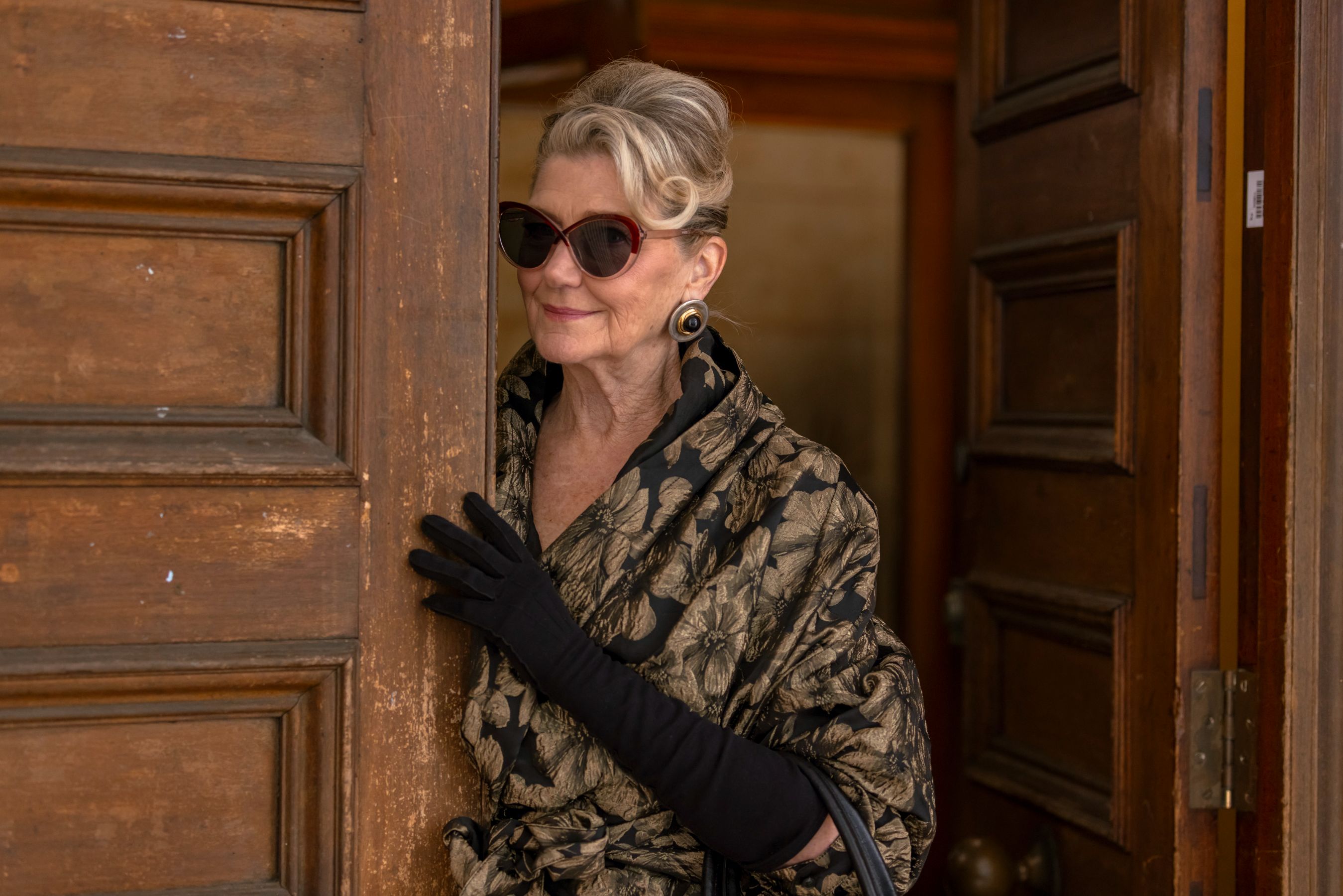 A stylish older woman stands partially behind a wooden door, wearing oversized sunglasses and elegant black gloves. Her hair is styled in soft waves, and she wears a floral-patterned wrap, exuding confidence and sophistication.
