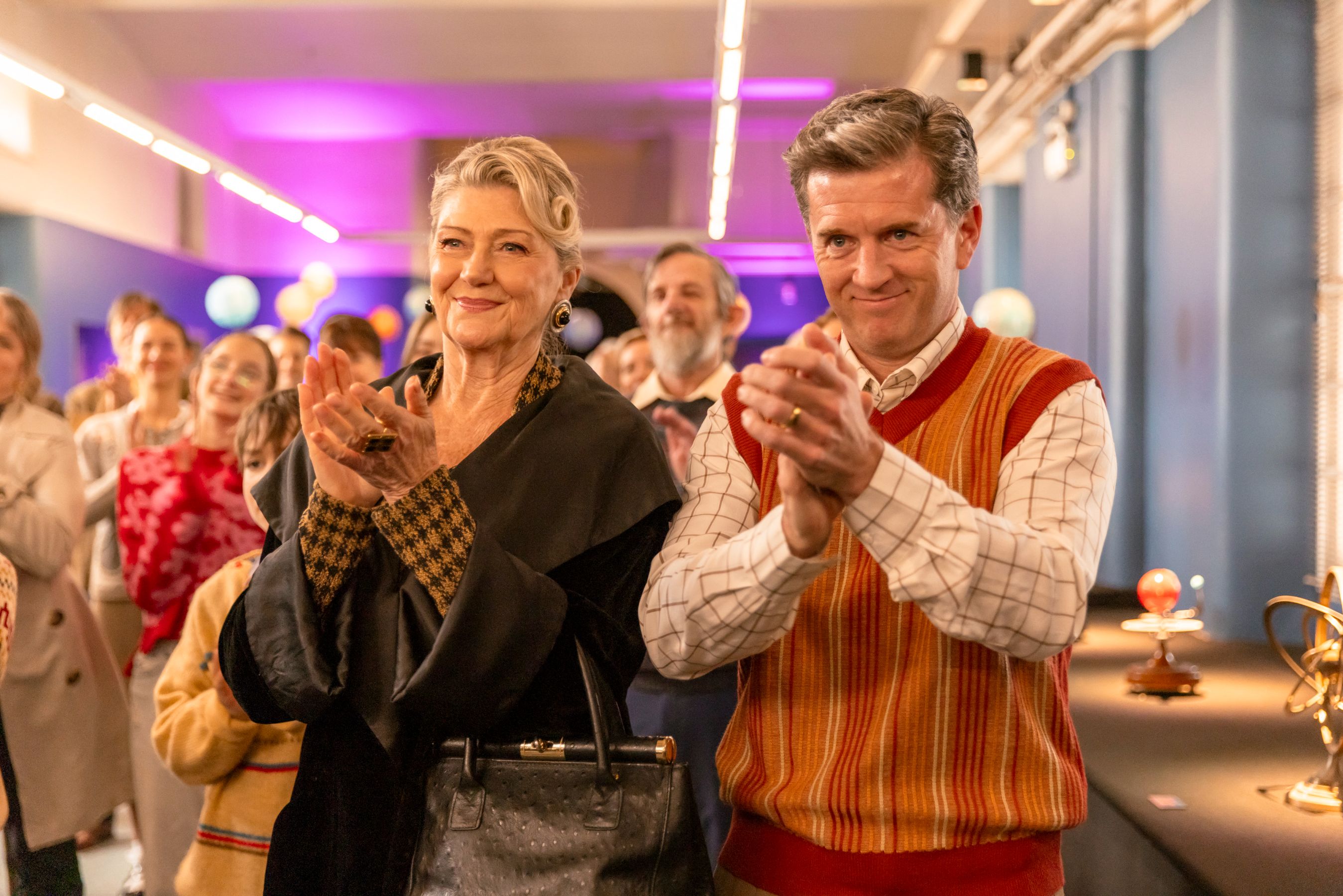 A woman with styled hair and elegant attire smiles while clapping her hands, standing beside a man in a colorful, retro sweater who also claps. They are joined by a crowd in a festive setting, with colorful lights illuminating the background. The atmosphere appears celebratory and joyful.