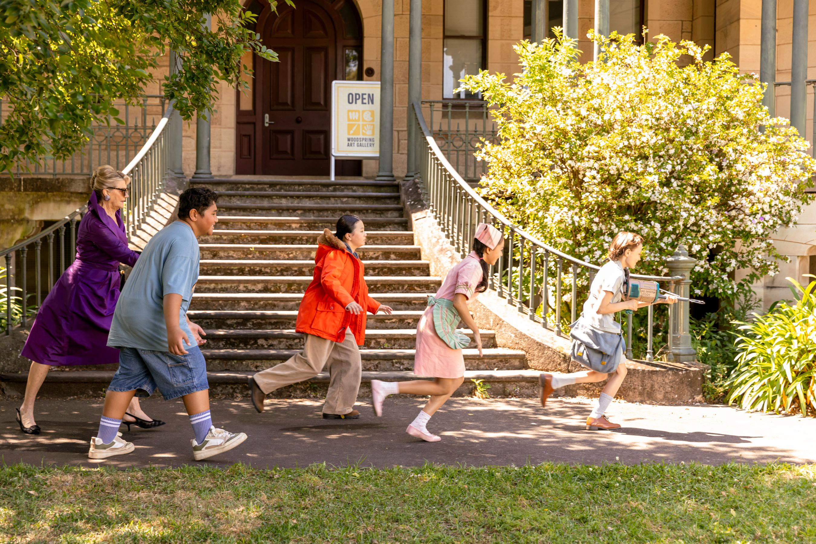 Five people are running down a pathway beside a set of stairs leading to a building. They appear to be in a playful or hurried chase, with colorful outfits including a purple dress, an orange coat, and a nurse's uniform. Lush greenery and flowering bushes surround them, creating a vibrant outdoor scene.