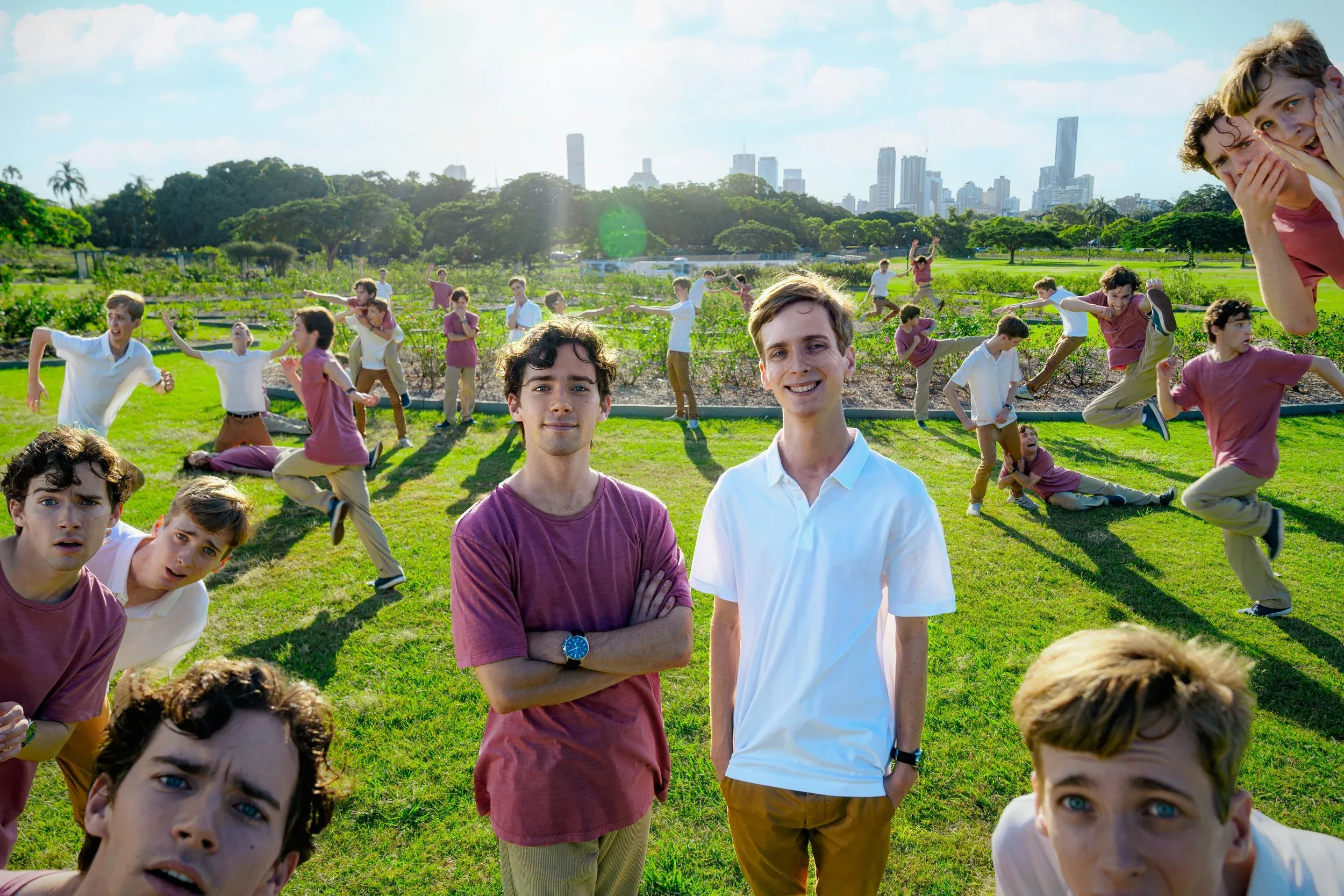 A group of young men is posing in a bright, sunny park, with some standing still and others in dynamic, playful poses, creating a sense of action and humor. In the background, the skyline of a city is visible against a blue sky, adding depth to the lively scene.