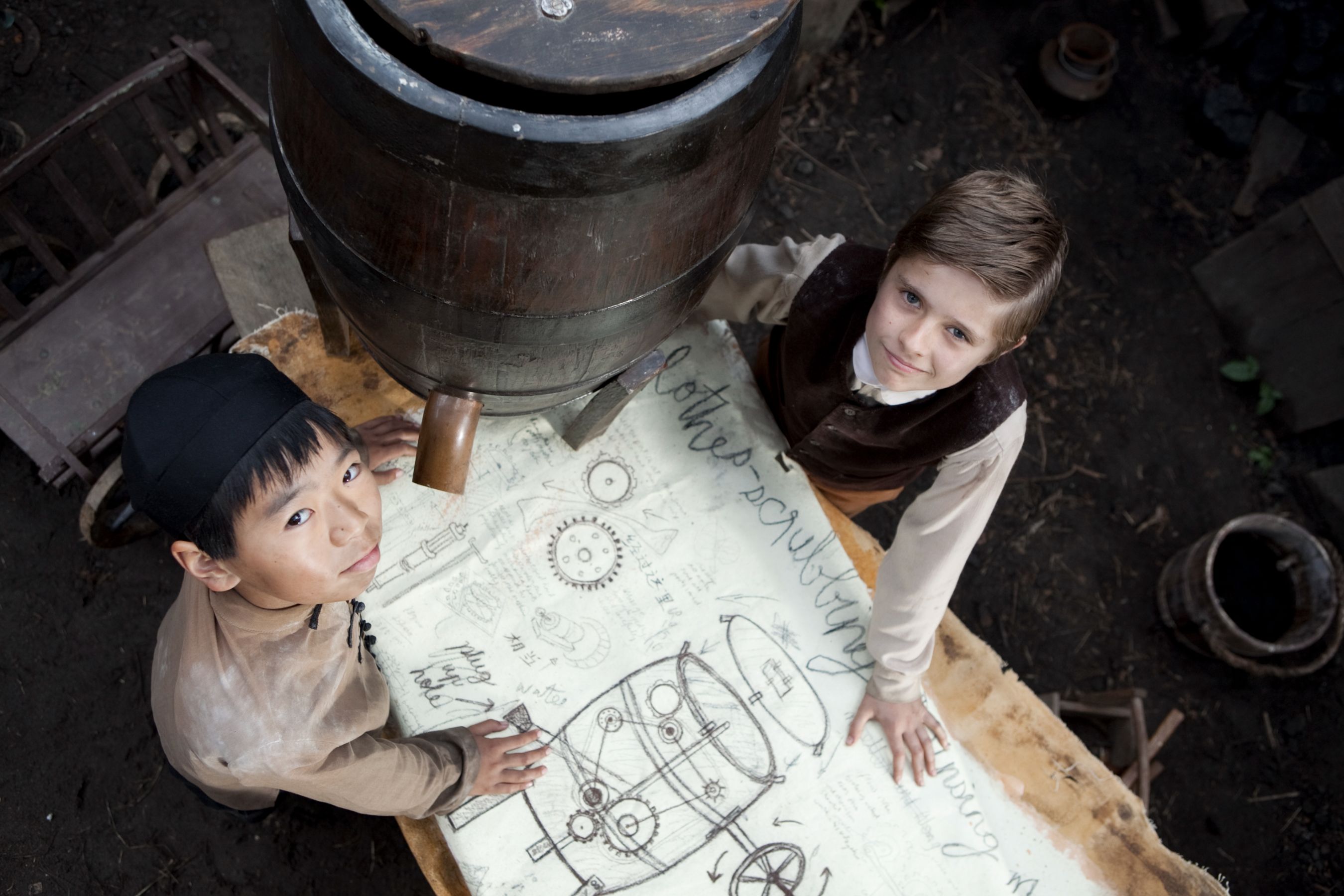 Two children are smiling as they stand next to a large wooden barrel, engaged in a creative project. They are positioned over a table covered with sketches and notes, including designs for machinery, written in a mix of handwriting and drawings. The setting appears industrial and rustic, with scattered tools and a dark earthen floor around them.