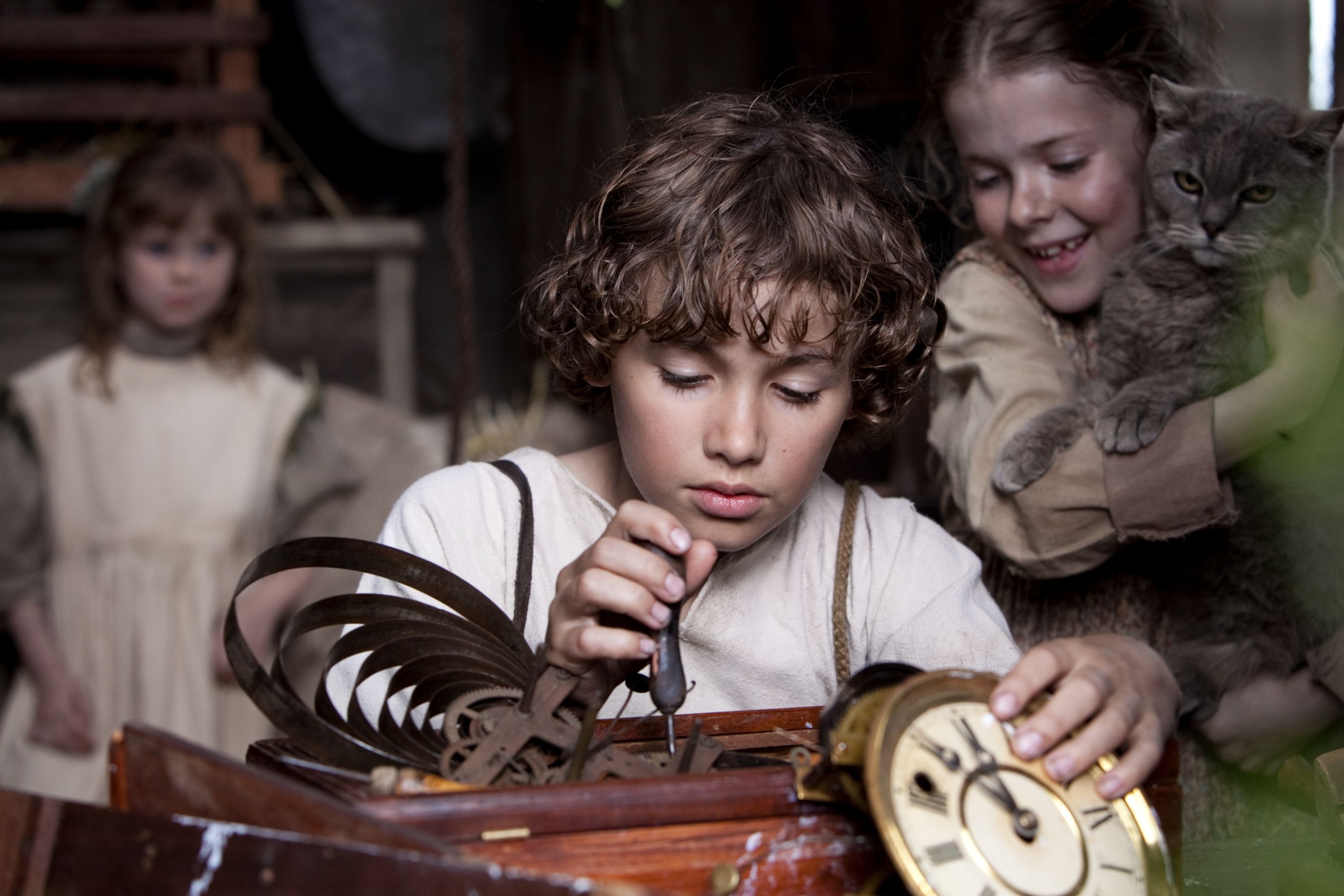 A young boy with curly hair examines a wooden clock mechanism, using a small tool with concentration. In the background, two girls, one holding a cat, watch him with smiles, creating a sense of camaraderie in the cozy, rustic setting.