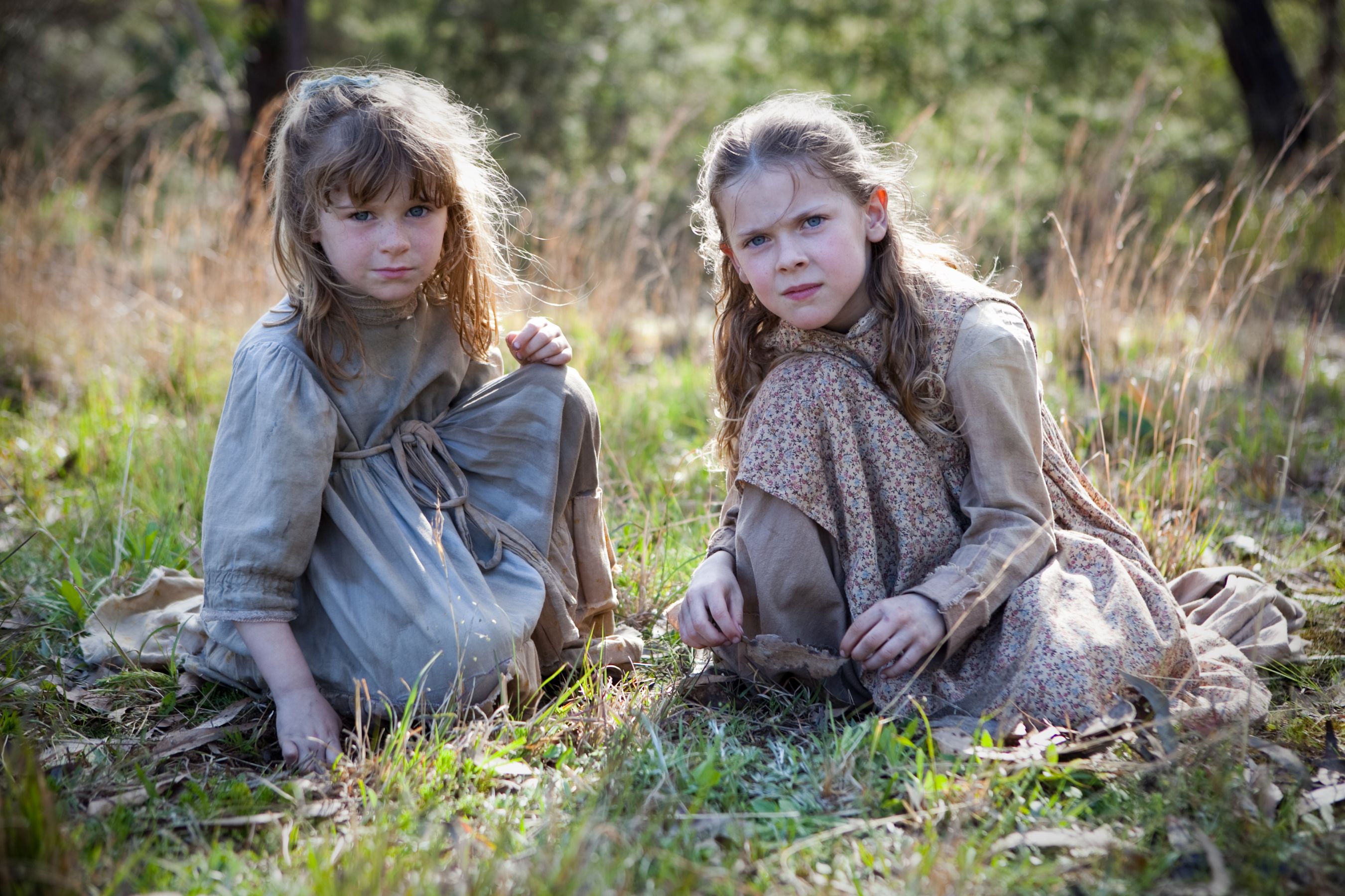 Two young girls sit in a grassy area, looking intently at the ground. One girl wears a simple gray dress and has tousled hair, while the other is dressed in a patterned brown outfit and gazes with a serious expression. Sunlight filters through the trees, illuminating their surroundings.