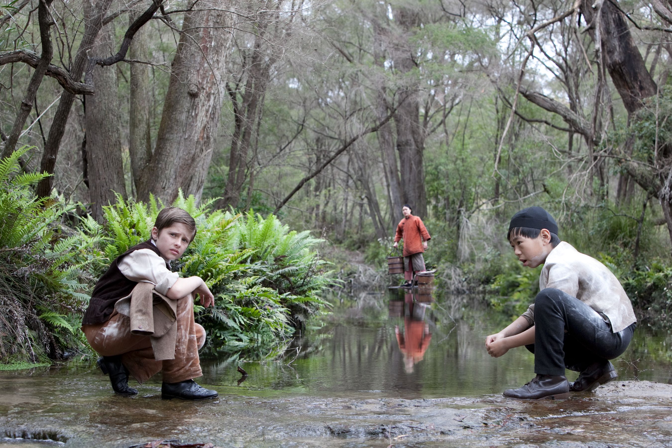 Two boys crouch by a calm stream in a forested area, one gazing thoughtfully at the water. In the background, a man stands on the bank, dressed in a bright orange shirt, partially obscured by leafy ferns and trees.