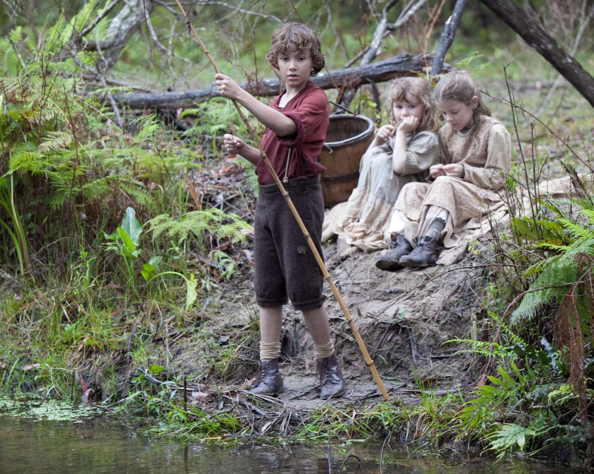 A young boy stands at the water's edge, holding a stick, while two girls sit quietly on the bank, engaged in their own activities. The scene is set in a lush, green environment with ferns and trees surrounding them.
