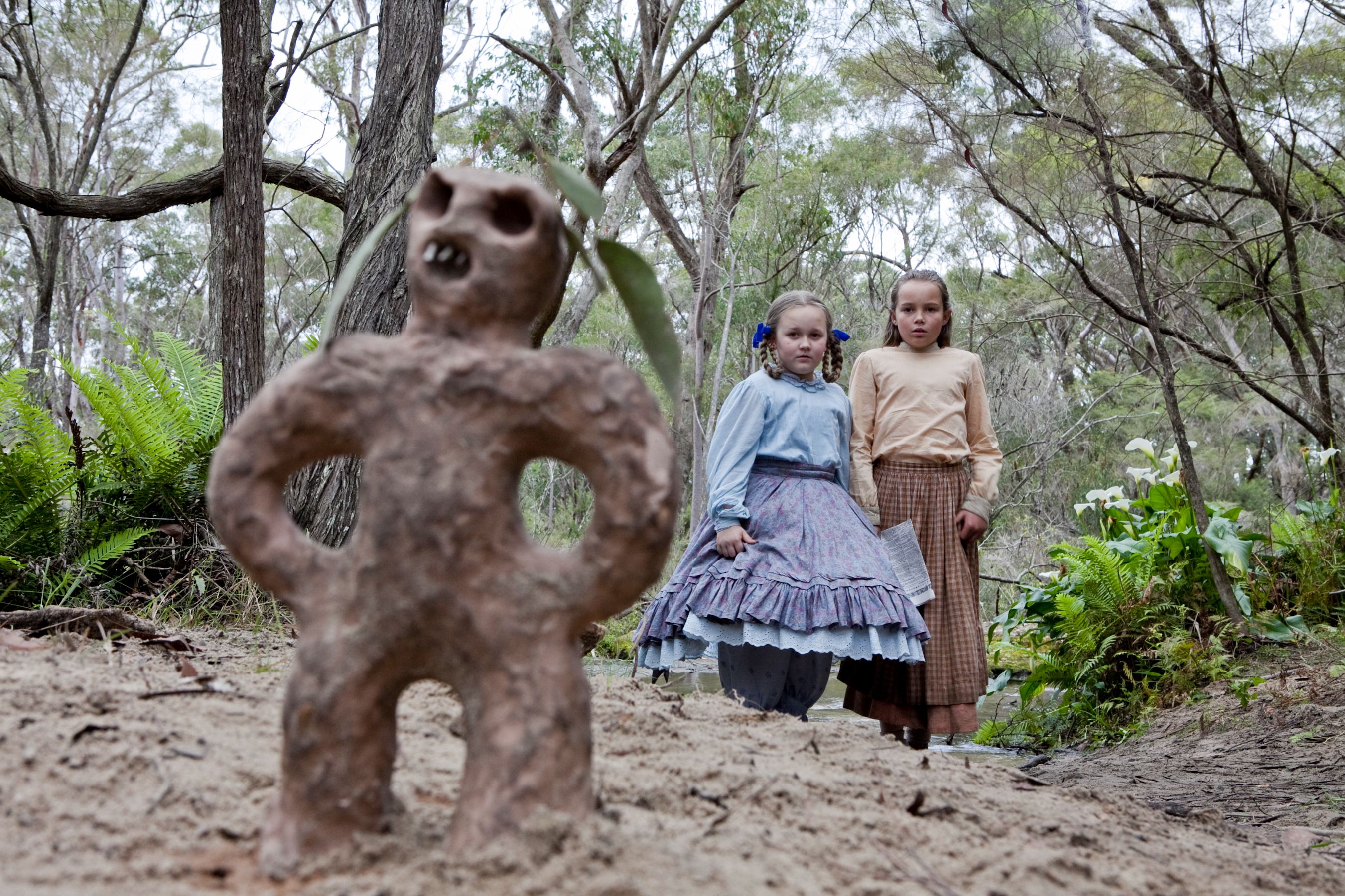 In a forest setting, a small, stylized clay figure with an aggressive facial expression stands prominently in the foreground, while two girls dressed in old-fashioned clothing watch curiously from a short distance. The background is filled with trees and greenery, creating a natural atmosphere.