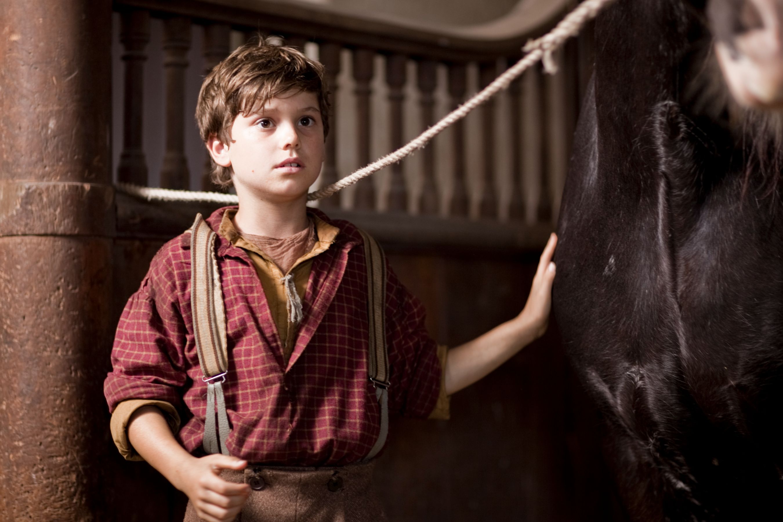 A young boy stands in a barn, looking up with a concerned expression as he gently touches the side of a large black horse. He is dressed in a plaid shirt and suspenders, with a rustic backdrop that includes wooden beams and a rope.
